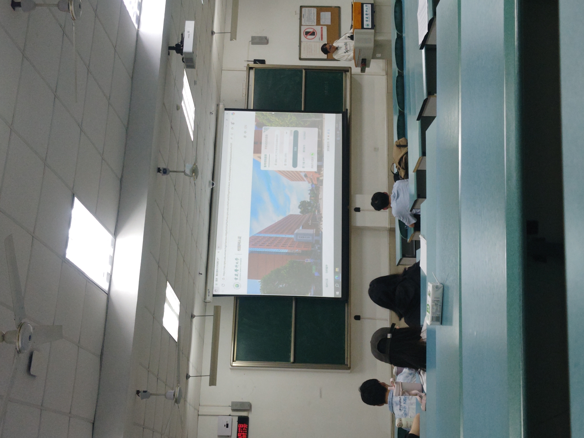 Here is a matter-of-fact description of the scene:
The image depicts a classroom setting in Chongqing High-tech Zone, China, likely at Chongqing Medical University, as indicated by the screen display. A professor stands at the front of the class near a screen and blackboard. Several students are seated at desks in the classroom. The classroom has standard features like ceiling lights, fans, and air conditioning units. A clock on the wall indicates the time. The environment appears academic, with students presumably engaged in a lecture or presentation.