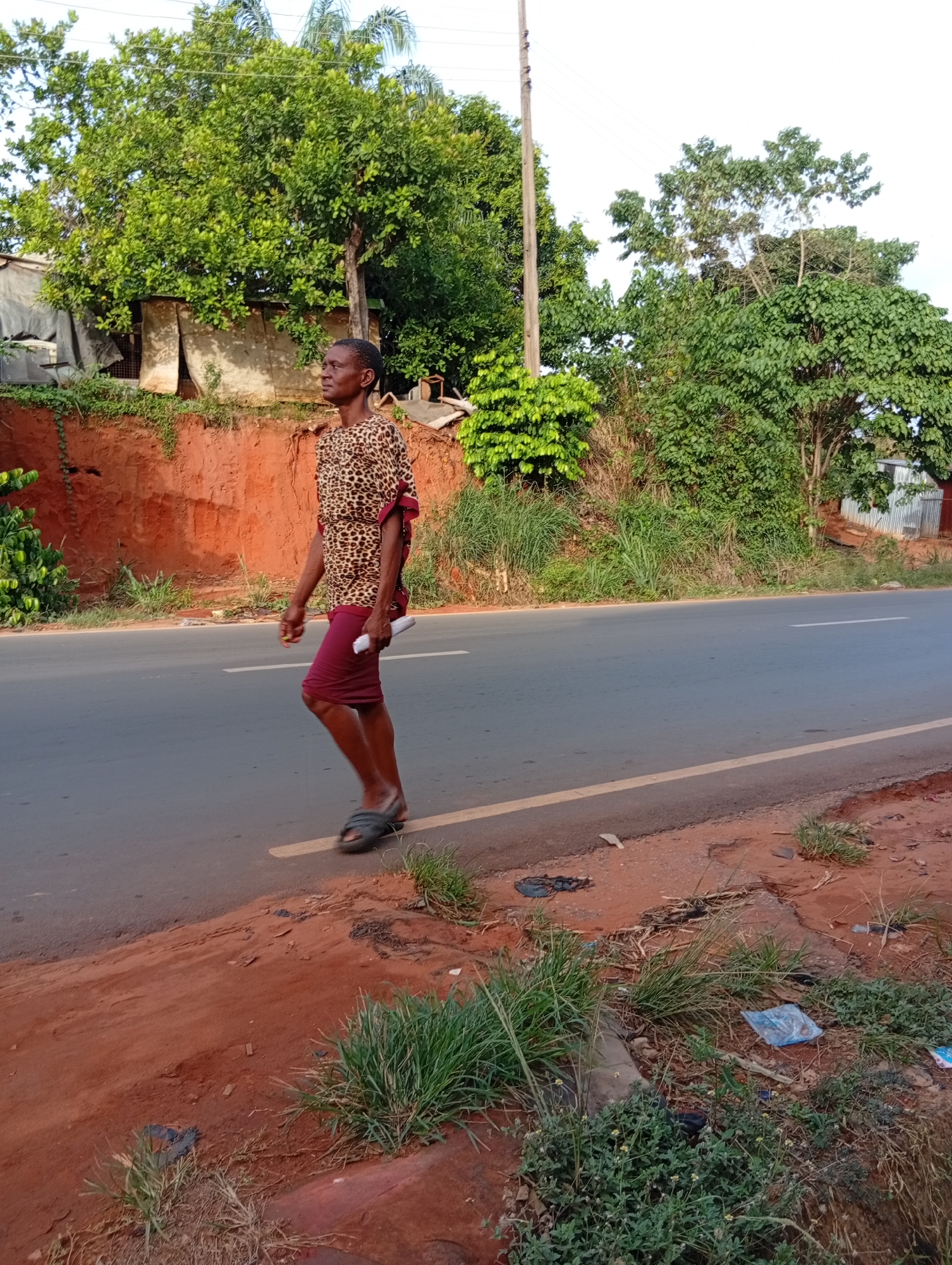 The image depicts a woman walking along the edge of a paved road in Nkpor, Nigeria. The road appears to be a two-lane road with a yellow dividing line and some litter near the edge of the road. The woman is walking away from the camera. She is wearing a leopard-print shirt and a dark skirt, and she is holding a rolled-up object. She is wearing sandals. The location has red soil on the roadside. The background consists of trees, vegetation, and buildings. A utility pole is visible.