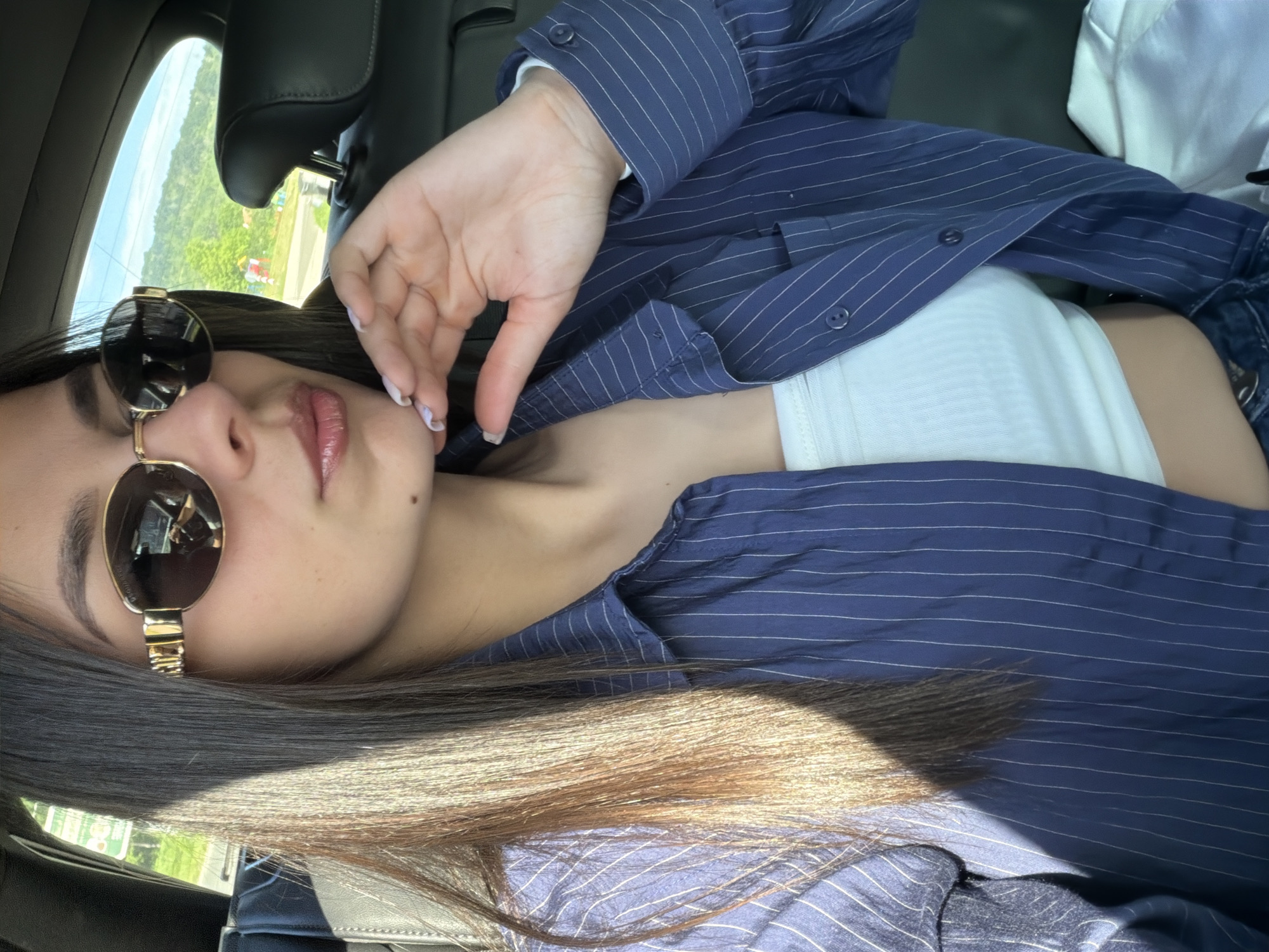 A young woman wearing sunglasses, a white tank top, and a blue pinstripe button-down shirt is in a car. The location is inferred to be Dryanovo, Bulgaria based on the query. The view from the car window is partially visible, showing greenery. The woman is posing with her hand near her face.