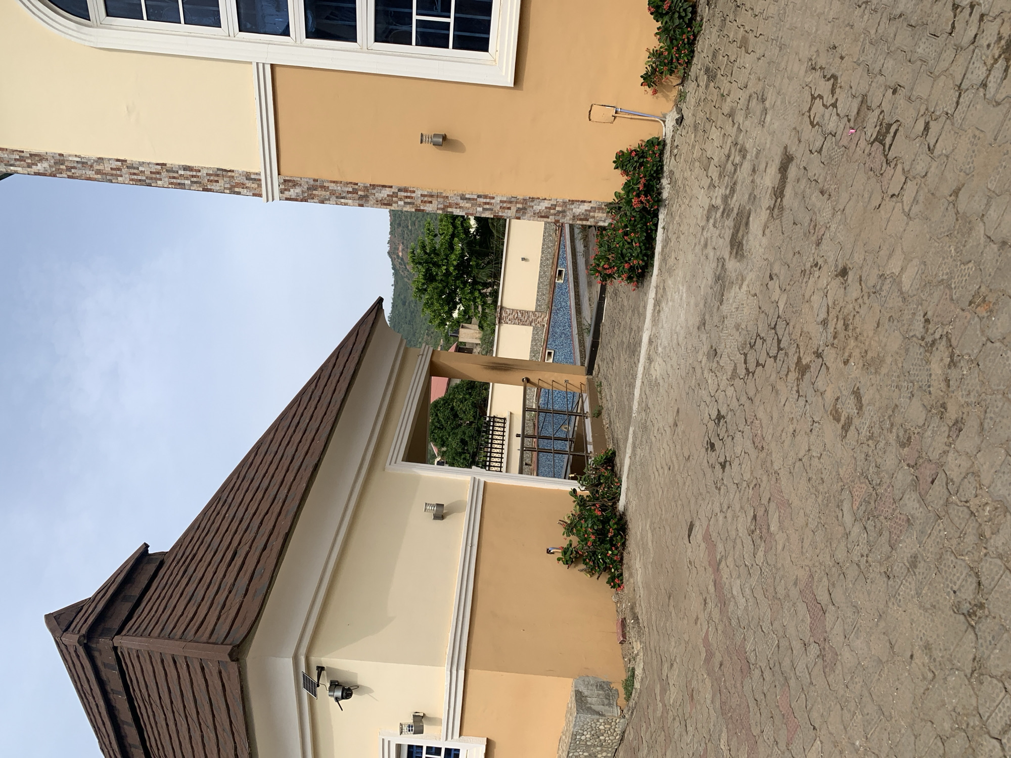The image shows a view of buildings and pavement in Lokoja, Nigeria. The architecture is modern residential, with features like a brown tiled roof, two-toned walls (cream and tan), and white-framed windows. There is a swimming pool visible between two buildings. A brick-patterned paved area is in the foreground. Red flowers are seen near the base of the buildings. The sky is overcast.
