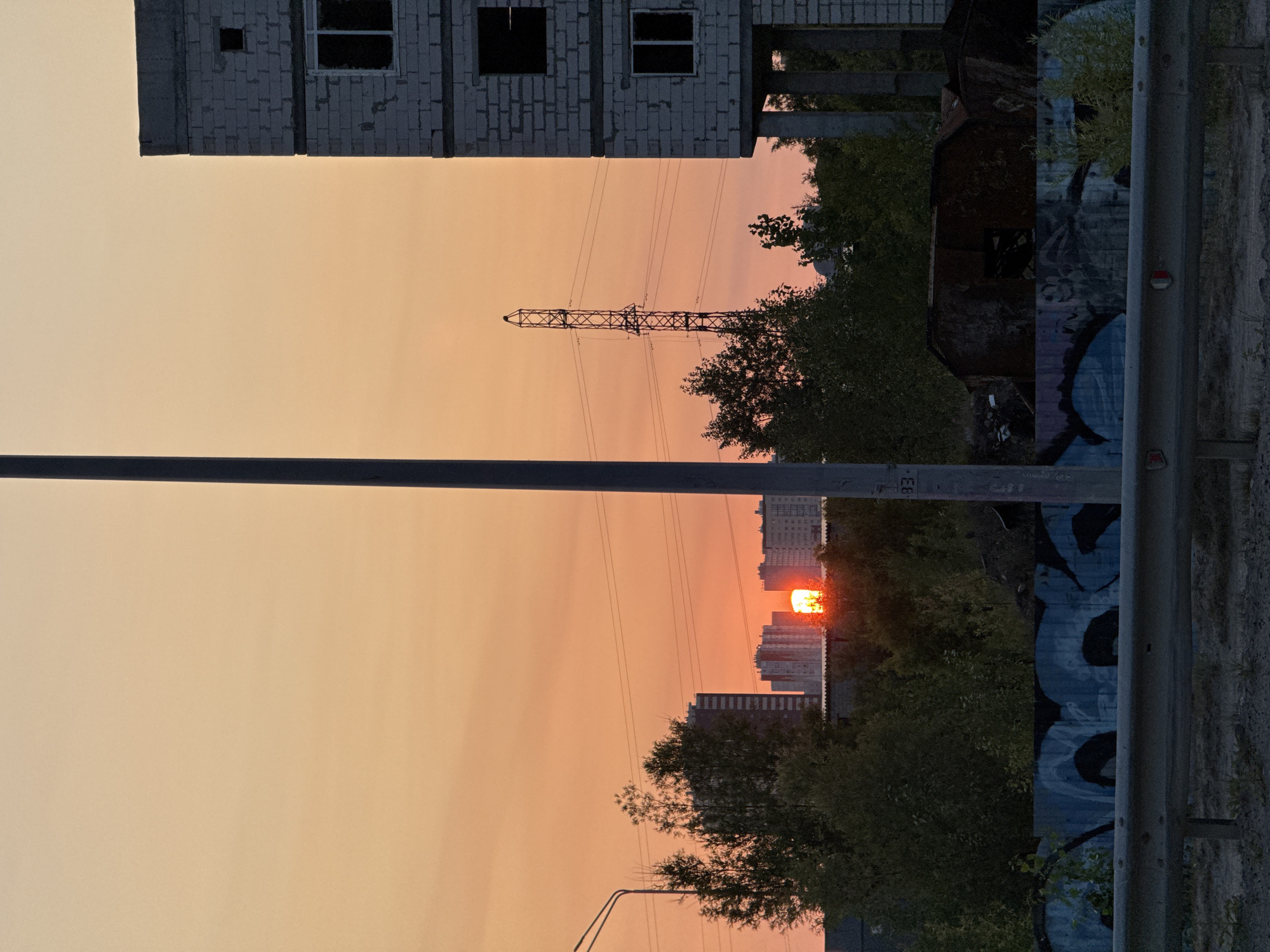 The image shows a sunset in Surgut, Russia. The sky is a gradient of orange and yellow. In the background, there are several tall buildings, including a high-rise under construction. A power line tower is visible to the left of the unfinished building. In the foreground, there is a metal guardrail and graffiti on a wall. The scene appears to be an urban or industrial area.
