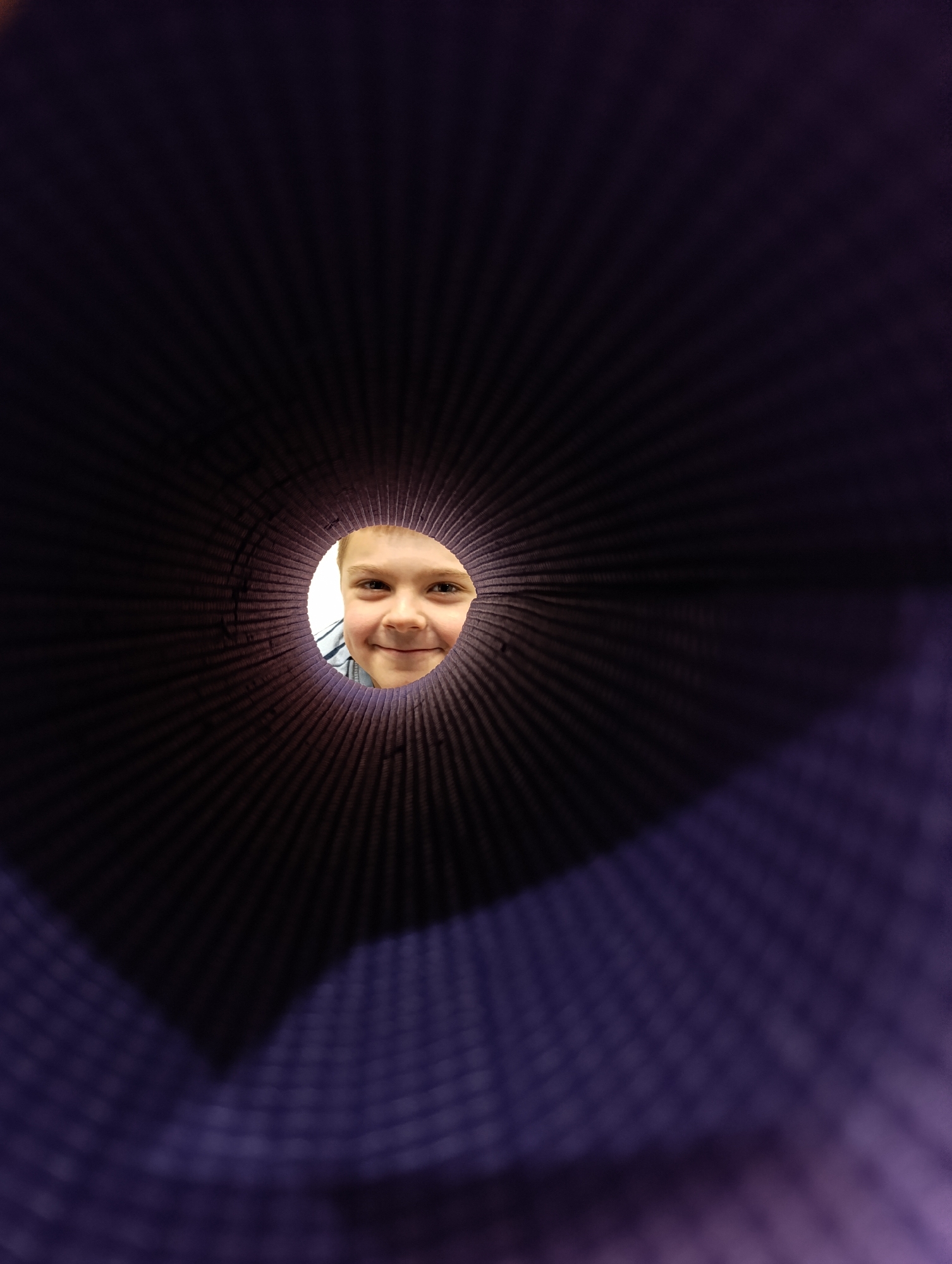 The scene, located in Chaikovsky, Russia, depicts a young child's face viewed through the circular opening of a rolled-up, dark purple, ribbed mat. The child, a light-skinned individual with light brown hair, smiles directly towards the viewer, with both eyes visible and a slight upward gaze. Part of a light-colored, possibly striped shirt is visible on the left side of the child's neck and shoulder. The interior of the rolled mat forms a dark, textured tunnel around the child's face, with radiating lines from the ribbed material. Beyond the child, through the aperture, a plain, brightly lit white surface is visible, consistent with an indoor wall. The majority of the image periphery is dominated by the dark purple, textured material of the mat, which appears out of focus in the foreground sections. The action captured is the child peering and smiling through the object.