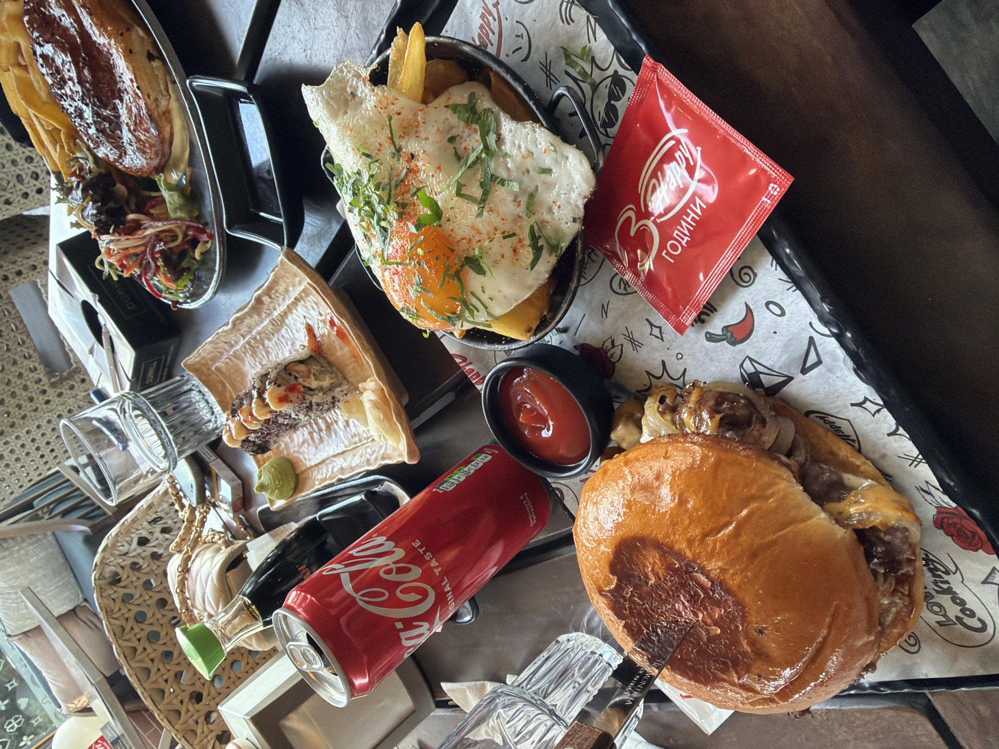 The image shows a table laden with food, including a sandwich, fries topped with a fried egg, what appears to be duck with vegetables, and a sushi-like dish. A can of Coca-Cola and a small dish of ketchup are also visible on the table. A packet of sauce labeled "Happy 3 Години" which translates to "Happy 3 Years" in Bulgarian indicates the location is likely in Varna, Bulgaria. A glass and a purse are also visible on the table. The scene suggests a meal being enjoyed at a restaurant or cafe.