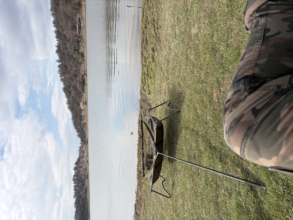 The image displays a first-person perspective of a lakeside scene in Cherna Gora, Bulgaria. In the immediate foreground, the knees and thighs of a person wearing camouflage-patterned trousers are visible. Directly ahead, on a patch of sparse green grass, rests a dark green fabric fishing cradle or unhooking mat, supported by a metallic four-legged frame, with an additional stabilizing pole extending towards the camera. A mesh net is visible within the cradle. To the right, near the water's edge, a green and yellow marker post with the numeral "5" is partially submerged. The mid-ground is dominated by a calm body of water, reflecting the sky and distant features. The far bank is lined with a density of bare deciduous trees or shrubs, suggesting a cooler season. Scattered along the distant bank are a small white structure, a larger building with a blue roof, and a wooden gazebo. The sky above is partially cloudy with patches of blue visible through white and grey clouds.