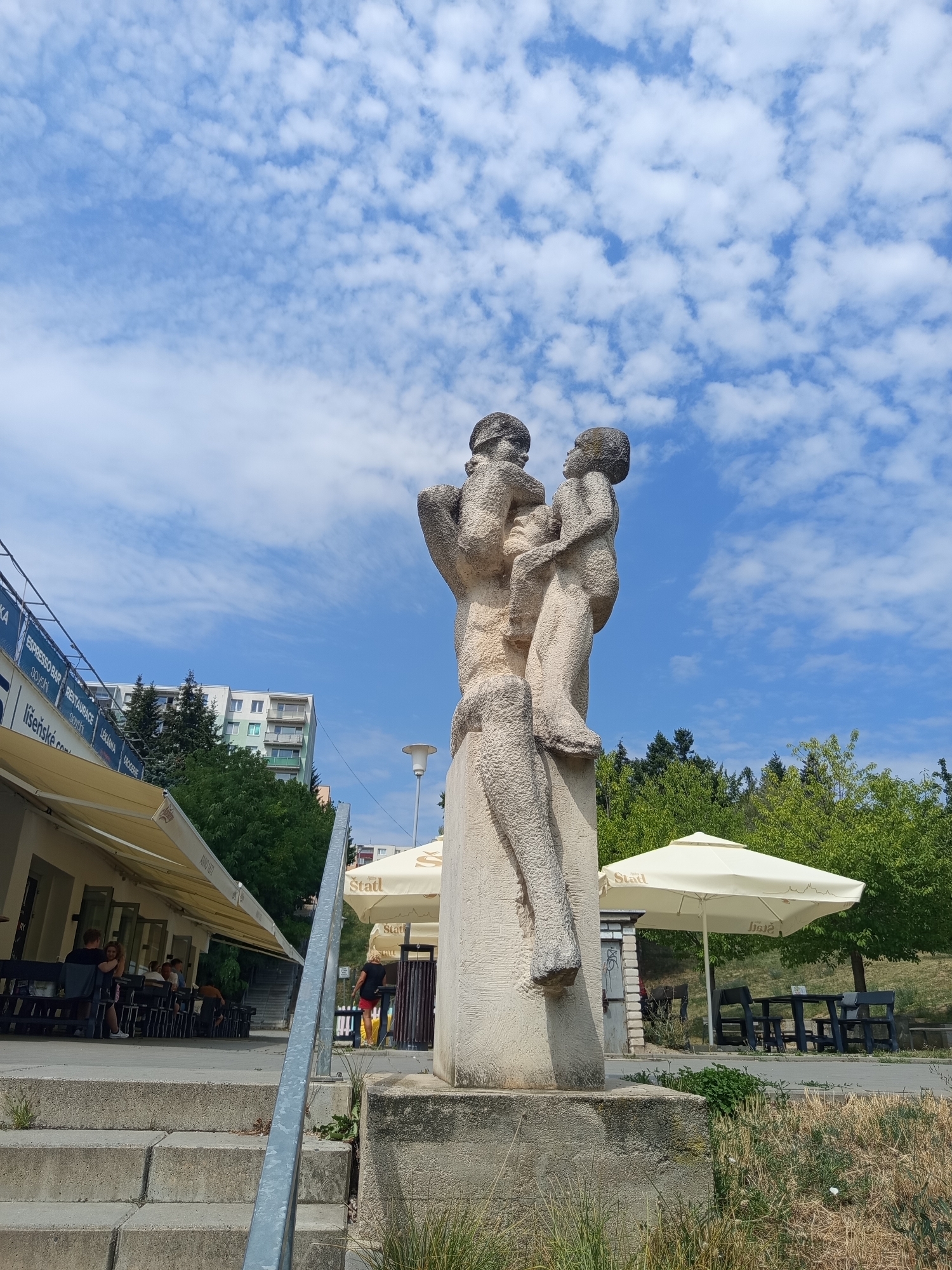A stone sculpture depicting two figures, possibly children, stands on a pedestal against a bright blue sky dotted with white, puffy clouds. The figures are rendered in a rough, textured style. To the left of the sculpture, a building with awnings and outdoor seating suggests a cafe or restaurant. People are seated at tables under the awnings. Further in the background, a multi-story residential building is visible, along with trees. To the right of the sculpture, outdoor seating with umbrellas is set up, and a woman in a black shirt and red shorts walks away from the viewer. The scene appears to be an outdoor public space, possibly a park or plaza, with elements of urban architecture and greenery. This sculpture is located at Lužánecký park in Brno, Czechia, specifically near the "Beseda" cultural center.