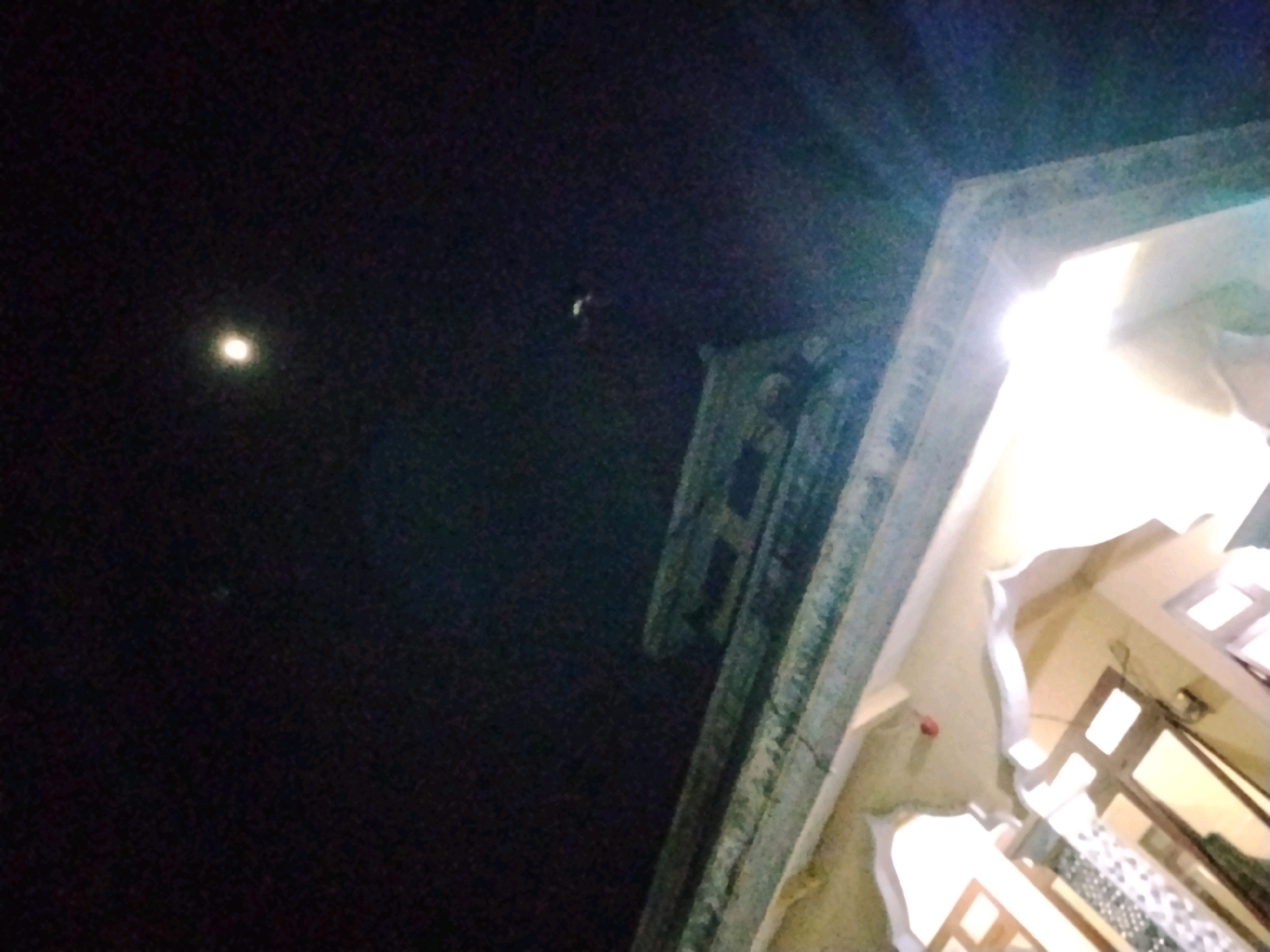 A low-light, angled shot captures a view of a moonlit sky and a building. The sky is dark, with a bright, out-of-focus circle representing the moon visible in the upper left quadrant. A faint, small, unidentified object appears in the sky to the right of the moon. The right side of the image is dominated by a building with a large, bright light source shining from an opening, suggesting an interior is illuminated. Architectural details of the building, including an arched opening and window frames, are visible. The overall scene suggests nighttime in a possibly urban or semi-urban environment, with the building possibly located in Panggungan, Indonesia, as indicated. There are no people or discernible actions depicted.