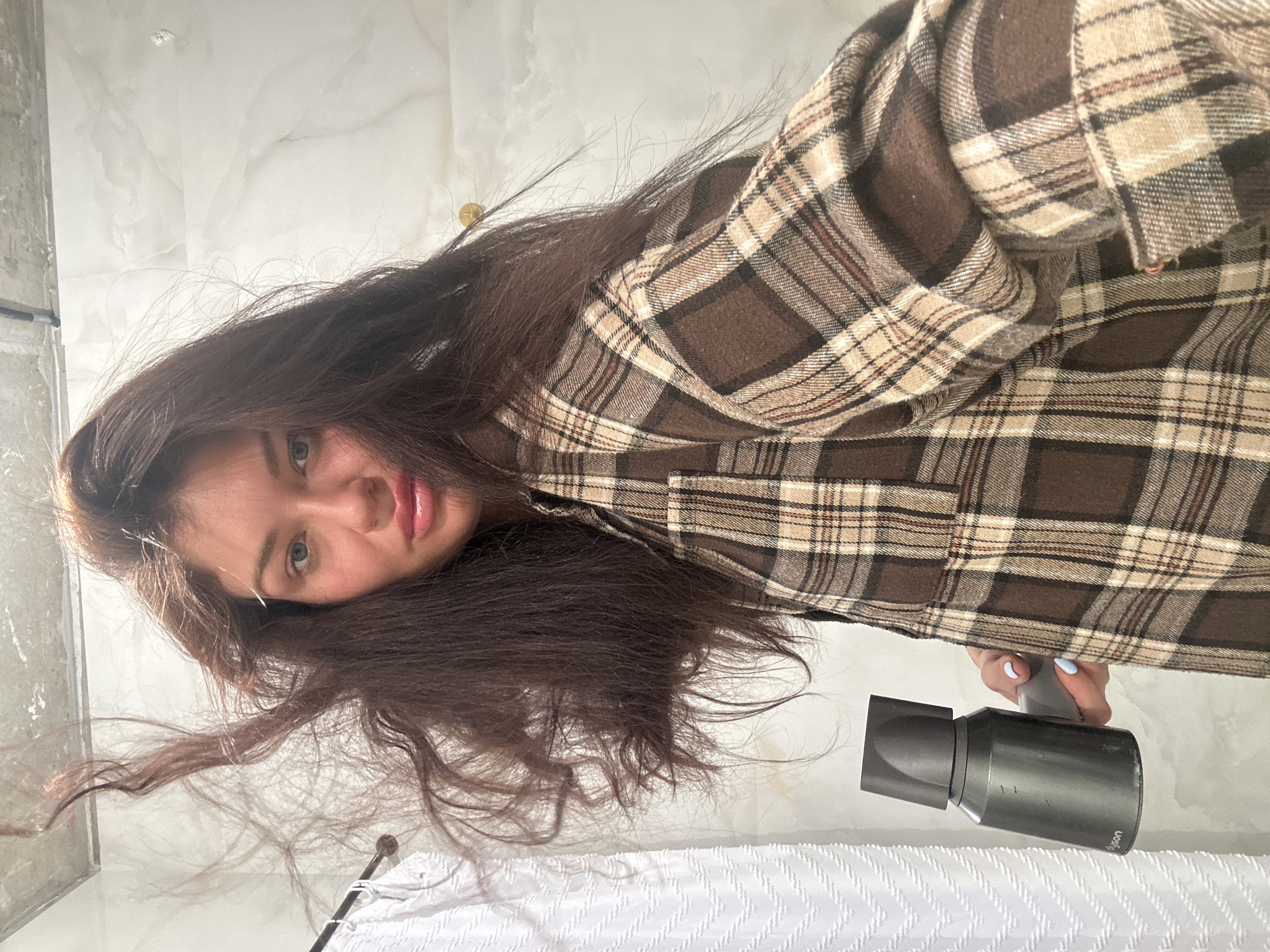 A woman with long brown hair is holding a grey Dyson hairdryer. She is wearing a brown and tan plaid shirt. The background appears to be a bathroom with white tiled walls. The location is not possible to determine from this image.