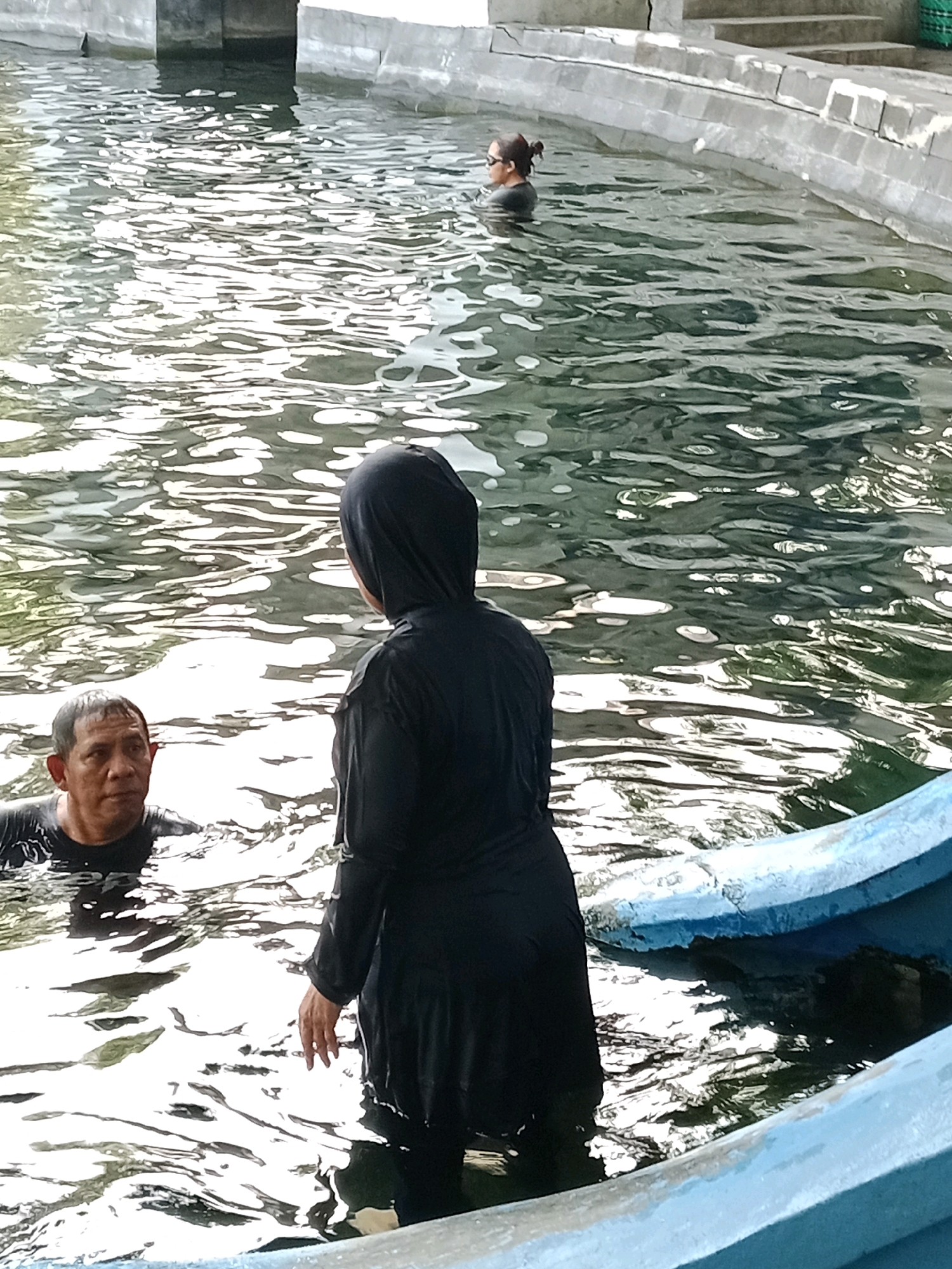 The image shows a partially filled outdoor swimming pool at a location identified as Jentekan, Indonesia. The water is rippling, with sunlight reflecting off the surface. In the foreground, a person wearing a black hijab and a full-length black swimsuit stands in the water, facing away from the camera. To the left of this person, a man with short, dark hair is partially submerged in the water, looking towards the person in the hijab. In the background, a woman with her hair in a bun and wearing sunglasses is swimming in the pool. A light blue boat or raft is visible in the lower right corner of the frame, partially filled with water. The edge of the pool is made of concrete or stone.