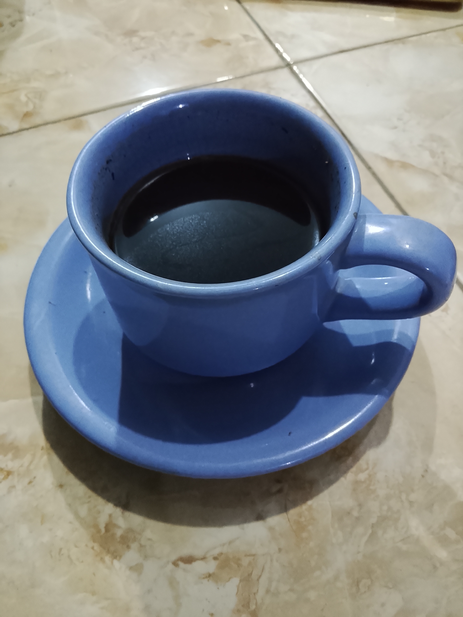 A blue ceramic cup filled with dark liquid, likely coffee, sits on a matching saucer. The cup and saucer are placed on a tiled surface with a marble-like pattern. The lighting suggests an indoor setting, possibly a home or cafe, with a shadow cast by the cup and saucer onto the tiles. The scene appears to be in Dawuan, Indonesia. There are no people or actions depicted.