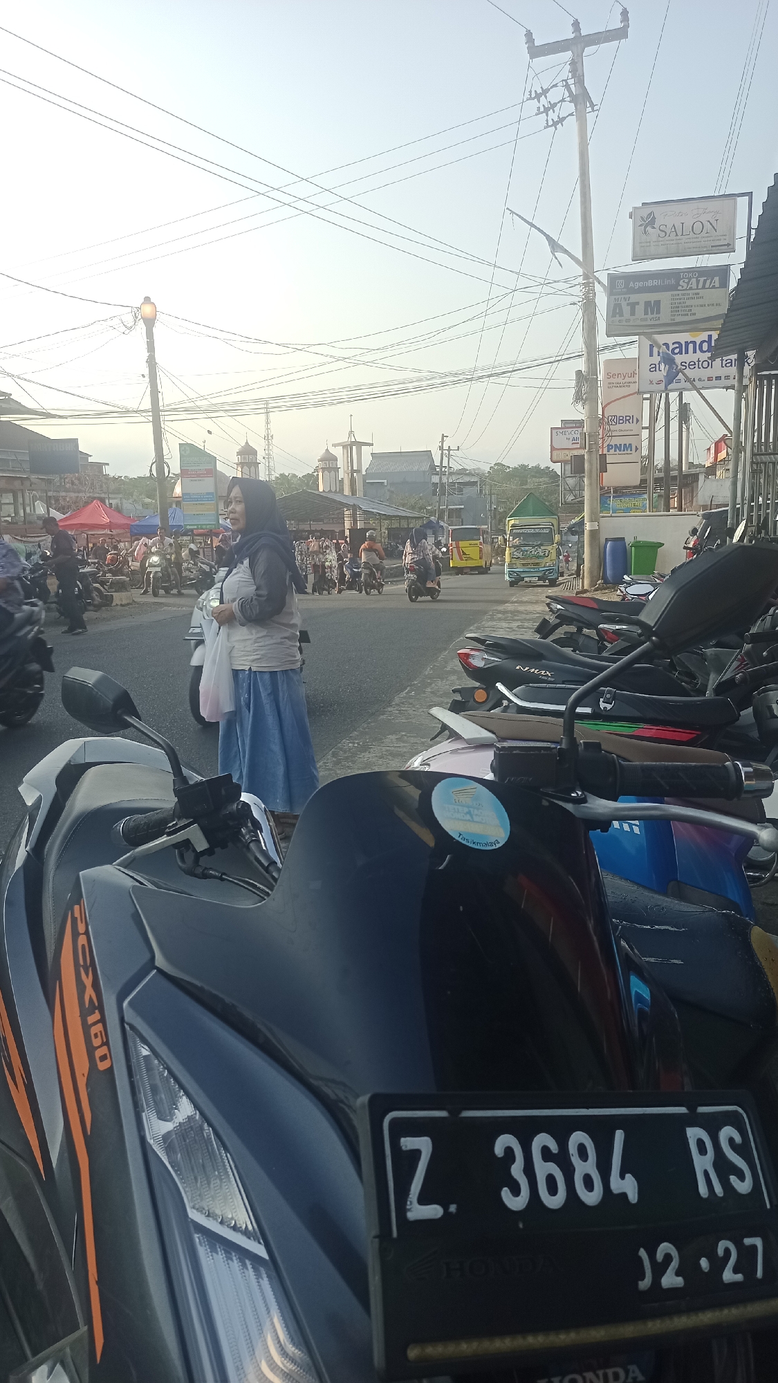 The image depicts a street scene in Karyamandala, Indonesia, likely during the late afternoon due to the warm, low-angle sunlight casting long shadows. In the foreground, the front of a dark-colored scooter with an orange "PCX 160" decal is visible, along with its license plate which reads "Z. 3684 RS" and "02.27". Several other scooters are parked behind it, suggesting a busy area.
A woman wearing a grey long-sleeved top, a dark headscarf, and a light blue skirt stands in the middle ground, holding a white plastic bag. She appears to be looking to her left, towards the street.
The street is a two-way road with moderate traffic. To the left of the woman, there is a market or roadside stall area with red and yellow tents. Several people are milling about and on motorcycles. In the background, buildings, including structures with domes that resemble mosques, are visible. A yellow bus and a green truck are also present on the road.
On the right side of the street, a series of advertisements and signs are mounted on poles. These include signs for "SALON", "TOKO SATIA", "ATM", "mandiri", "SENYUM ALL", and "PNM". The overall environment suggests a commercial and residential area in a town or city. The sky is clear with a pale blue hue, and a network of electrical wires crisscrosses the upper portion of the image.