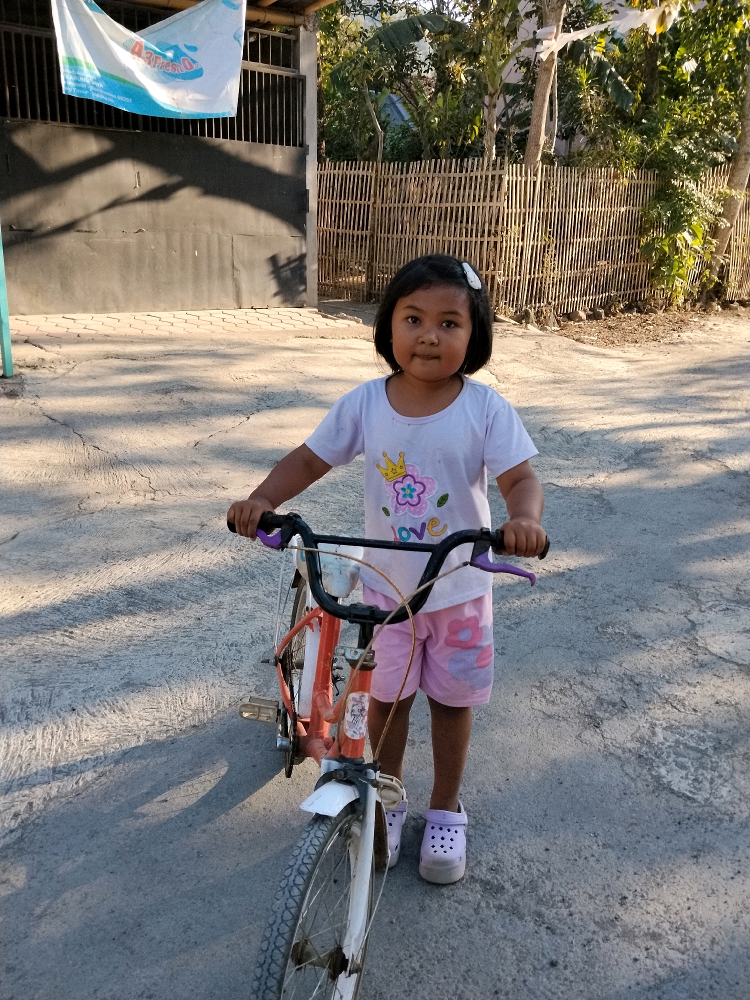 A young girl stands on a cracked concrete surface, holding the handlebars of a small orange bicycle. She is wearing a white t-shirt with a floral design and pink shorts with floral accents. Her feet are clad in light purple clogs. The bicycle has a white fender on the front wheel. Behind her, a bamboo fence borders a property, and a dark metal gate with vertical bars is visible. A white banner with blue lettering hangs above the gate. The scene is outdoors, with dappled sunlight casting shadows on the ground, suggesting it is daytime. The overall setting appears to be a residential area, possibly in Tlogosari, Indonesia, given the architectural style of the fence and gate.