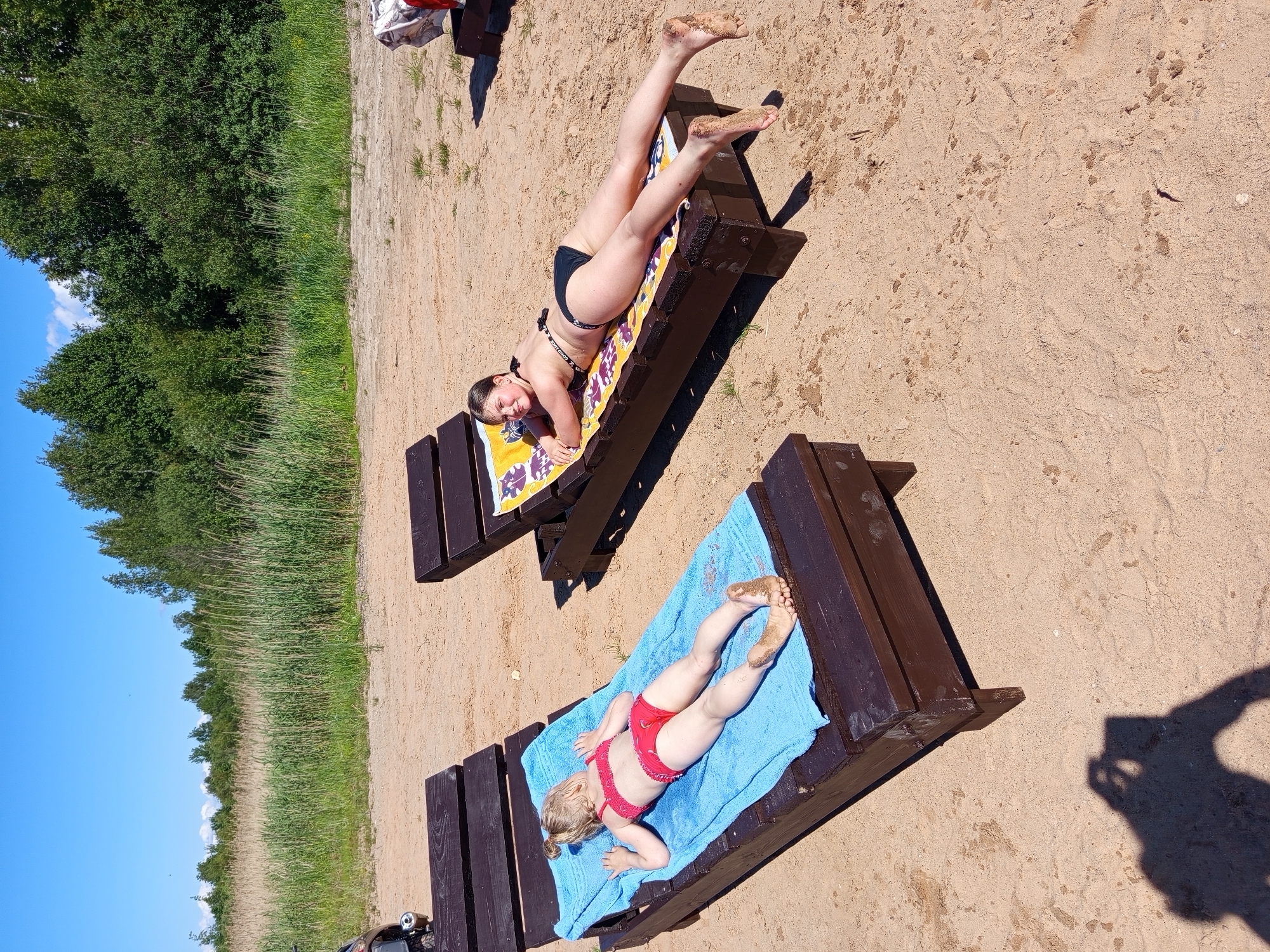The image displays an outdoor scene on a sandy expanse, consistent with a beach or lakeside environment, under a clear blue sky. Two individuals, both appearing female, are present, each lying prone on a dark brown wooden sun lounger. The subject on the left, a younger girl with light-colored hair styled in an updo, is positioned on a lounger covered with a light blue towel and wears a red two-piece swimsuit. Her bare feet, coated with sand, are visible. The subject on the right, an older girl or young woman with dark hair, is positioned on a lounger covered with a yellow and purple patterned towel and wears a black two-piece swimsuit. She is observed looking towards the camera with a subtle smile; her bare feet are also visible and covered in sand. In the lower left corner, the distinct shadow of a person holding a mobile device is cast onto the sand, indicating the photographer's position. The background consists of a dense strip of tall green vegetation, including what appear to be reeds or grasses, followed by a line of mature trees. The specific location within the city of Ostrovtsy, Russia, is not discernible from the visual information provided.