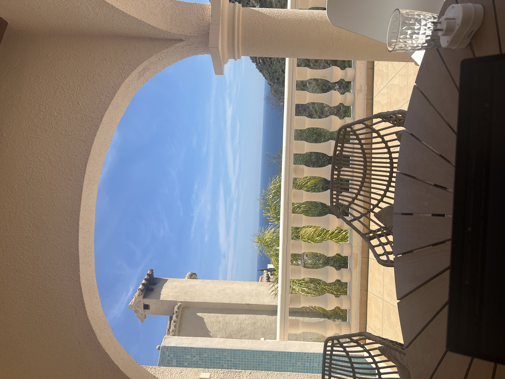 The image shows a view from an arched opening, possibly a balcony or veranda, with a table and chairs in the foreground. The sky is blue with some clouds. The sea is visible in the distance. A white balustrade is present along the balcony edge, with greenery behind it. A building with a tiled roof and a light blue tiled wall segment is seen below. The location is likely a building overlooking the sea in el Poble Nou de Benitatxell / Benitachell, Spain.