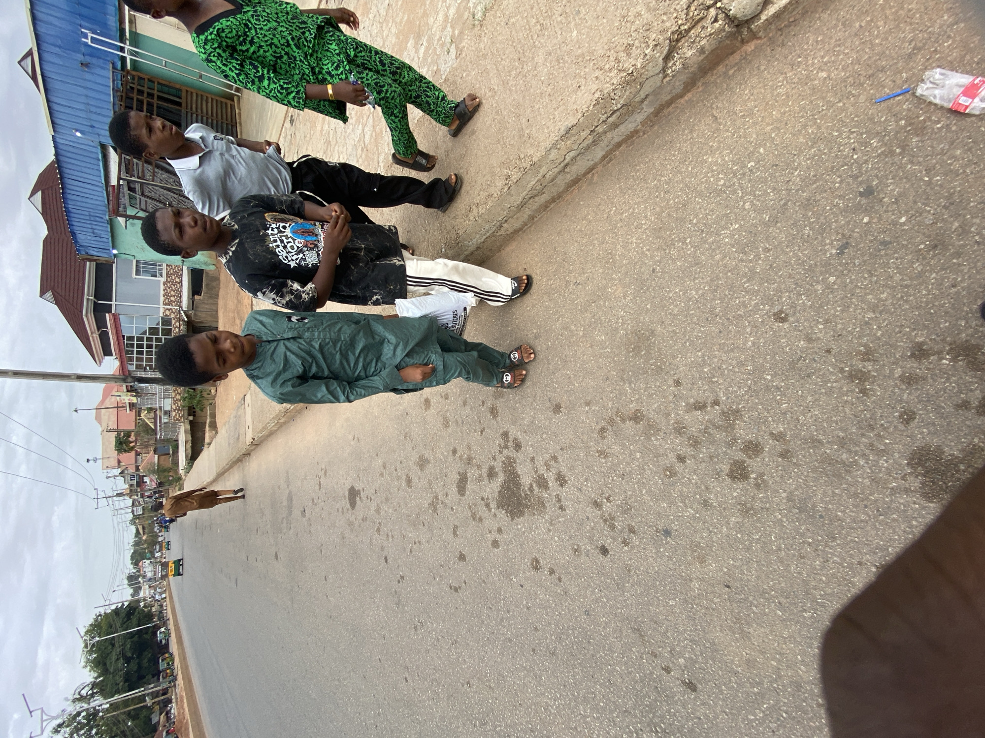 Four young males stand on the pavement in Bukuru, Nigeria. The location is on an asphalt road, with various buildings visible in the background. The young males are dressed in casual clothing. From left to right, the clothing consists of: a dark green matching set, a black graphic t-shirt with white striped pants, a gray shirt with black pants, and a green matching set. A brown dog is seen on the opposite side of the street. There is trash on the ground. Buildings are visible in the background of the scene. Power lines are stretched from building to building.