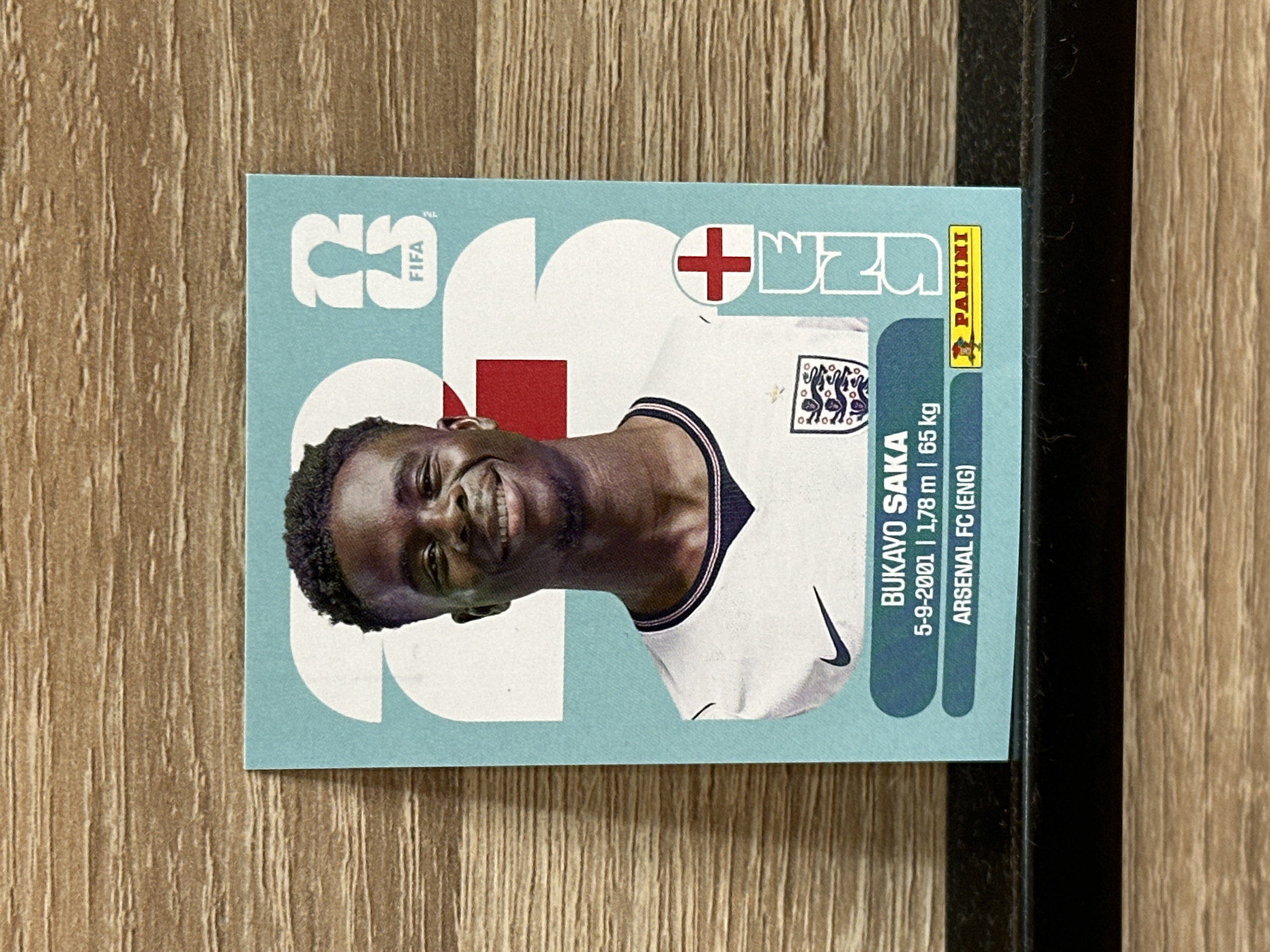 The image shows a close-up, eye-level, horizontal shot of a Bukayo Saka FIFA sticker. The sticker is placed on a wooden surface, possibly a shelf or table. The sticker features a portrait of Bukayo Saka wearing an England football kit, with the FIFA logo and the Panini logo visible. The sticker also displays his name, date of birth, height, weight, and club. The background is a light blue color with large white numbers and shapes. The overall lighting is bright and even. There is no information in the image or its context to suggest any connection to Prisovo, Bulgaria.