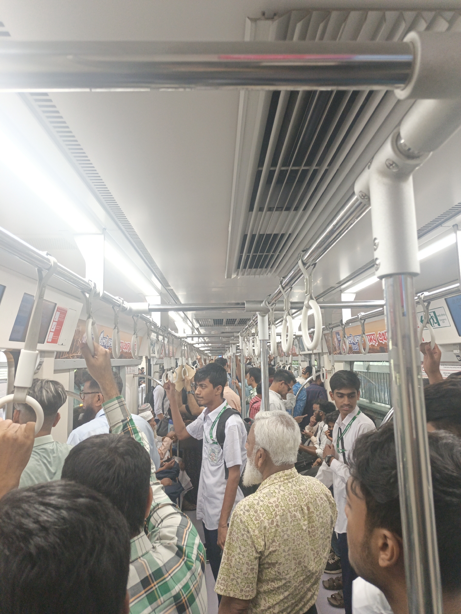 The image depicts a crowded metro train in Dhaka, Bangladesh. The interior is brightly lit with a modern design, featuring white and silver fixtures. Numerous passengers are standing and holding onto overhead handrails and loops. The crowd consists of men and women of various ages, dressed in casual attire. Some individuals are wearing what appears to be a student uniform. The overall impression is a typical scene of public transportation during peak hours in a densely populated city.