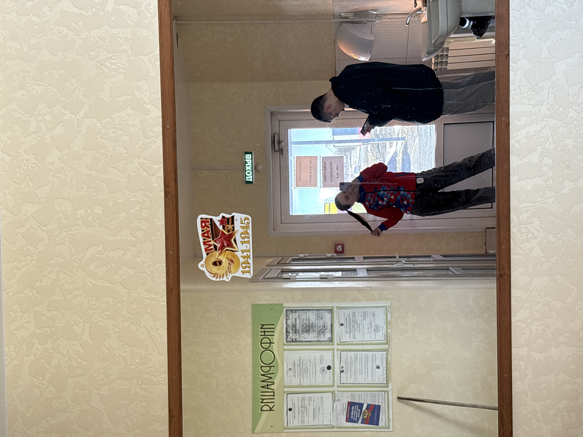 The image is a reflection in a mirror. The mirror is hanging on a wall with textured beige wallpaper. The wall also has a poster with the Cyrillic text "РПИАМДОФИН" on a green background and a collection of certificates. Above the poster, there is a sticker commemorating the years 1941-1945.
In the reflection, two individuals are visible. One is wearing a dark jacket and grey pants, looking at a phone. The other is wearing a red jacket and dark pants, also holding a phone. Behind them is an exit sign labeled "ВЫХОД" in Cyrillic, and a door. Beyond the door, there is a view of the outside, possibly a cityscape. The reflection also includes a white sink, a towel dispenser and ceiling lights. The overall scene appears to be an indoor space, possibly a hallway of a building. Since it is not possible to identify the location as the city of Gory, Russia from this image, there is no way to tell whether it is there.