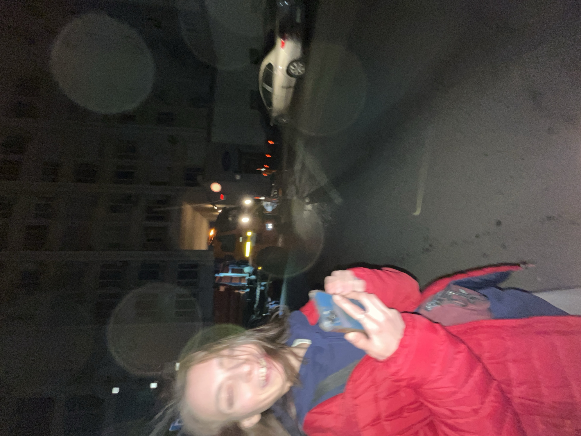 The image depicts a nighttime scene in an urban environment. A person, likely female, is holding a blue smartphone and smiling, clad in a red jacket and navy blue shirt. The background shows a city street with a parked white car, streetlights, and a building with illuminated windows. The image appears to be taken at an angle, causing a distorted perspective.
