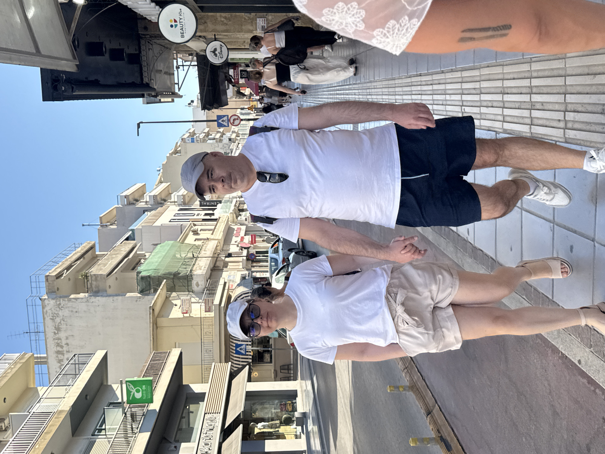 In Chania, Greece, a couple walks hand-in-hand on a sunlit sidewalk alongside a street lined with buildings. The man, wearing a grey baseball cap, white t-shirt, and dark blue shorts, walks towards the viewer, his gaze directed forward. The woman, beside him, wears a white t-shirt, light beige shorts, and beige sandals. She also looks ahead. Behind them, other pedestrians are visible on the sidewalk, and cars are parked and moving on the street. The buildings flanking the street appear to be commercial establishments, with shops and signs visible. The architecture suggests a typical Mediterranean urban setting.