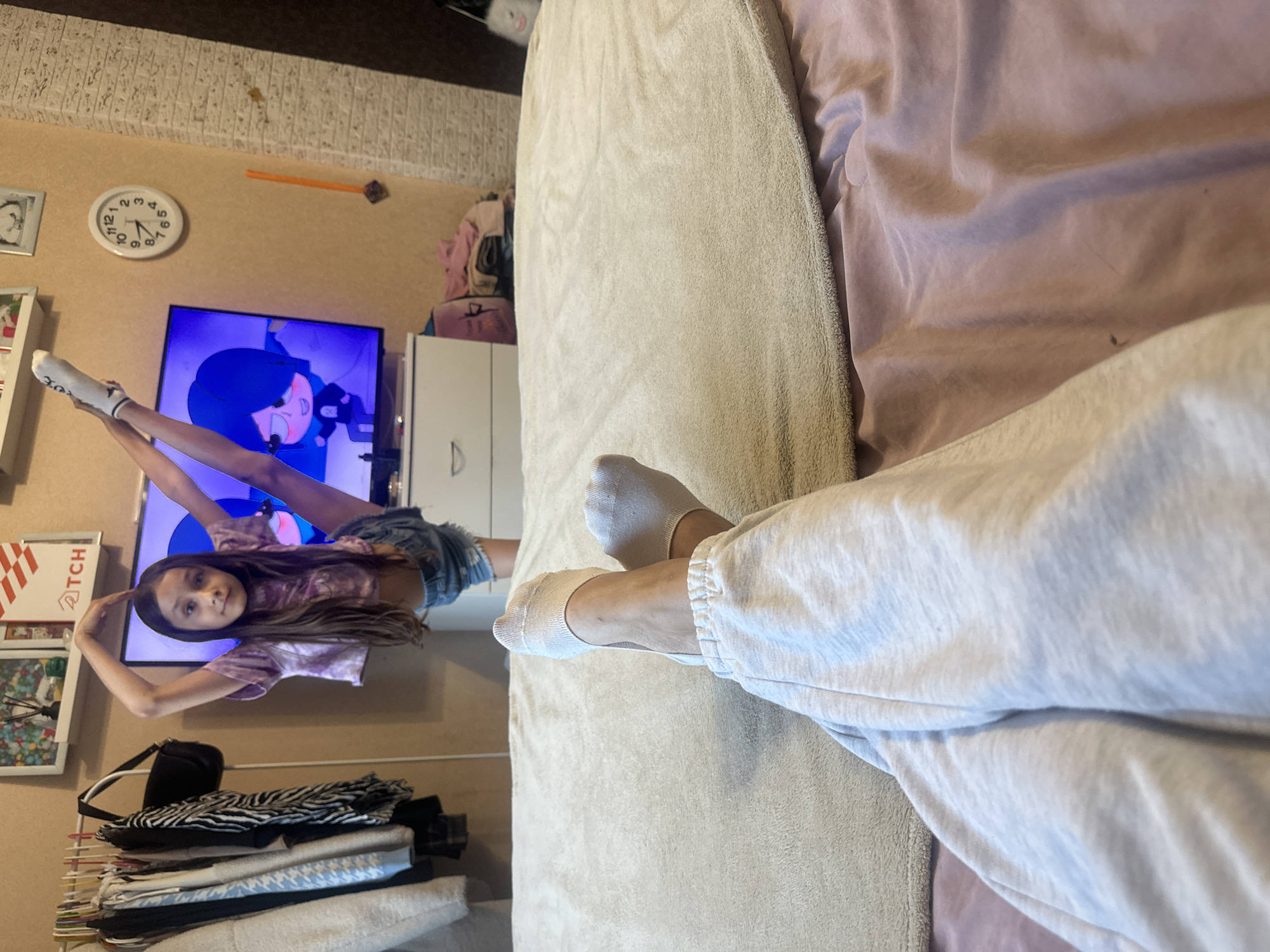 The media file depicts a young girl in a room, seemingly in a casual or playful moment. She is lying on her back on what appears to be a bed or sofa, with her legs extended upwards. She is wearing a light purple tie-dye t-shirt and denim shorts, with white socks on her feet. Her hair is long and dark brown, and she is looking directly at the camera with a neutral expression.
In the background, a television screen is visible, displaying an animated character with blue hair. To the left of the television, a round wall clock shows the time to be approximately 3:50. There are also some framed pictures on the wall, a white cabinet, and a stack of clothes on a rack. The overall setting suggests a domestic environment, possibly a bedroom or living room.
The lighting appears to be indoor artificial light, and there are no clear indicators of the time of day or weather conditions outside. The mood seems relaxed and informal, with the girl posing for the camera. The image is oriented vertically.
Visible text includes "TCH" on a red and white box to the left of the television and numbers on the clock face.
