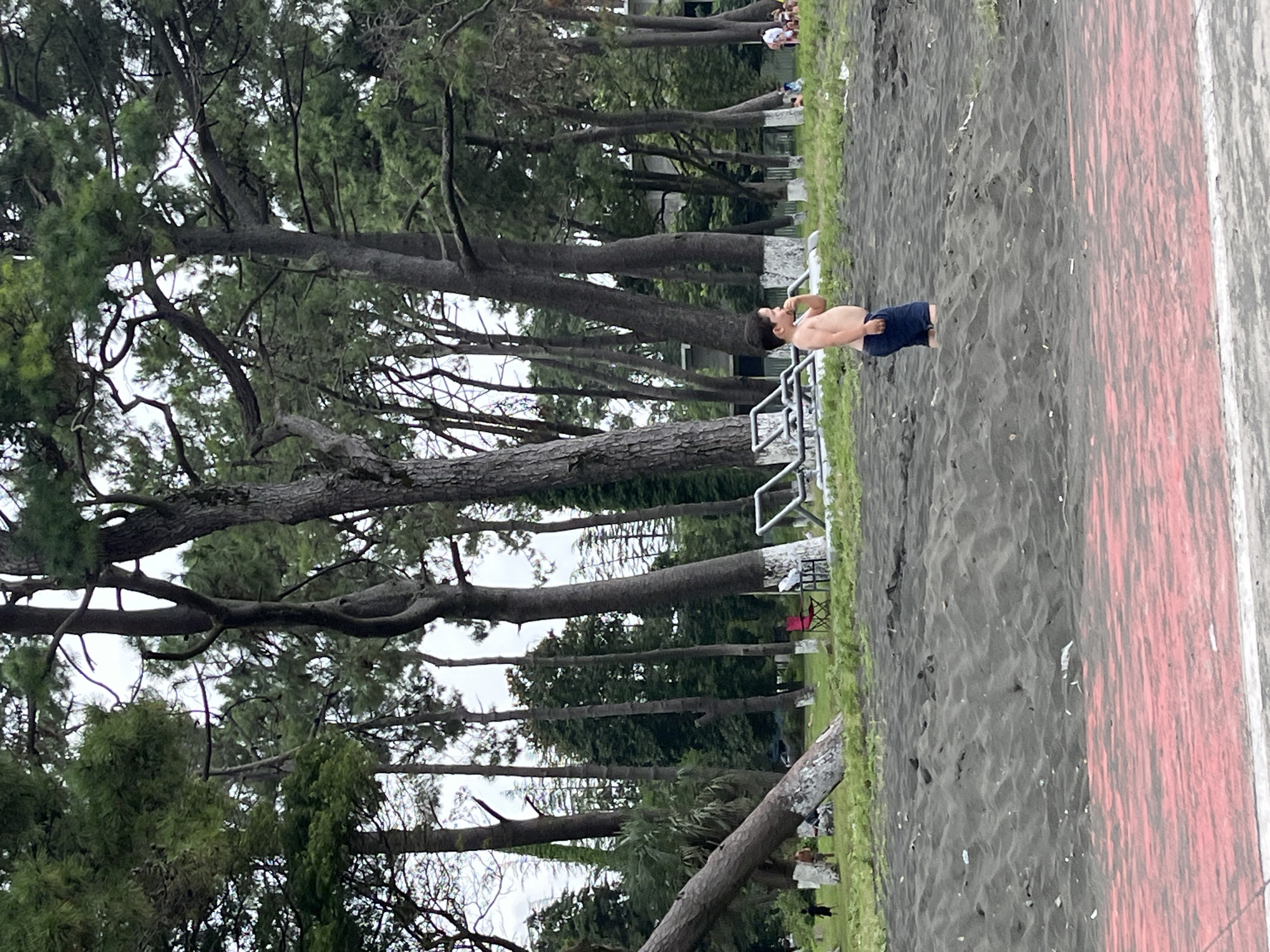 A young boy stands on a black sand beach, wearing dark blue shorts. He is facing right and appears to be looking at something off-camera. The sand is uneven, with small mounds and depressions. In the background, a line of tall pine trees with white-painted bases separates the beach from a grassy area. Several white structures, possibly part of a playground or park, are visible among the trees. Further in the distance, a few people are scattered across the grass, some sitting and others standing. The sky is overcast, suggesting a cloudy day. The foreground shows a strip of pinkish-red surface, possibly a paved walkway or a recreational area. The overall scene depicts a natural beach environment within a park setting in Ureki, Georgia, a town known for its magnetic black sand beaches.