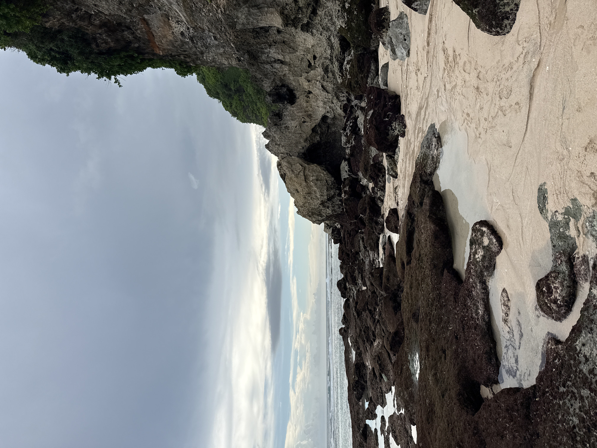 The image depicts a coastal scene in Ungasan, Indonesia. A rocky cliff covered with vegetation forms the upper left of the frame. Below the cliff is a sandy beach with scattered rocks and patches of shallow water. The sea stretches out towards the horizon under a cloudy sky.