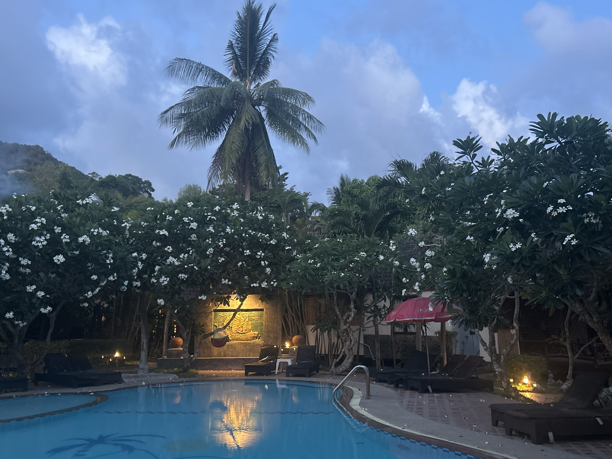 The image captures a serene nighttime scene at a resort in Baan Wang Ta Kien, Thailand. A kidney-shaped swimming pool dominates the foreground, its water reflecting the ambient light. Around the pool's perimeter are several dark lounge chairs, some adorned with white frangipani blossoms. Mature frangipani trees with clusters of white flowers frame the scene, adding to the tropical ambiance. Behind the trees is a wall with a lit bas-relief carving, flanked by decorative lighting. A tall palm tree rises above the trees, its silhouette contrasting with the cloudy evening sky. A glimpse of a hillside with dense vegetation can be seen in the background. A red umbrella is visible on the right side of the image near the lounge chairs.