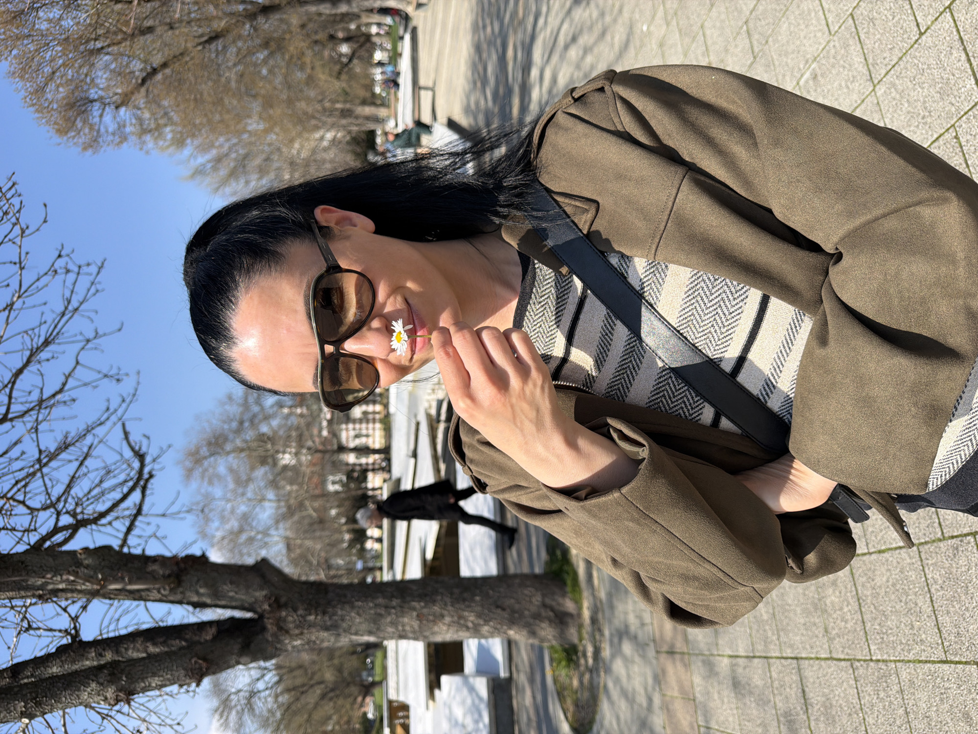 A woman with long dark hair, sunglasses, and a green jacket stands outside, holding a daisy near her nose. She is wearing a patterned top and a black shoulder bag. The background includes trees without leaves and a paved area. The city of Pleven, Bulgaria, can be inferred from the location, based on visual clues. A person in dark clothing is walking in the distance.