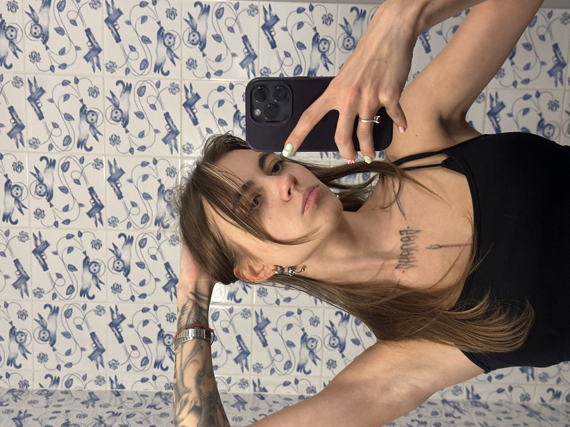 A young woman with long brown hair, wearing a black tank top, takes a mirror selfie in a bathroom. She has tattoos on her left arm and chest. Her nails are painted with a green and white design. She is wearing a ring on her right ring finger and a watch on her left wrist. The bathroom walls are covered in blue and white patterned tiles, featuring images of cats and guns. The woman is holding a black smartphone in her right hand. The lighting is natural, possibly from a window or overhead light. The image appears to have been taken in a residential bathroom. There is no information available to suggest the location is in Moscow, Russia.