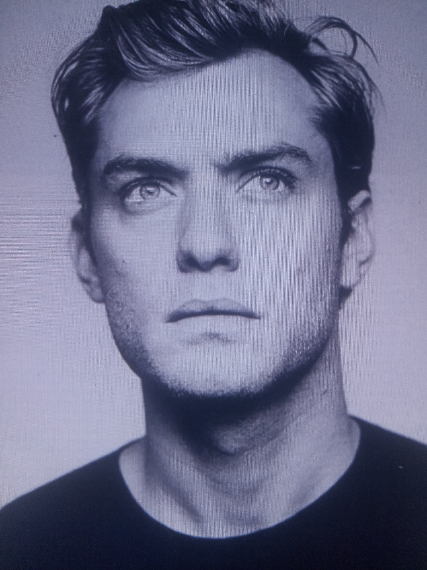 This is a black and white, head-and-shoulders portrait of a man. He has light-colored hair styled with some volume on top and to the side, and is looking upwards and to his right. His eyes are open and appear to be blue. He has a clean-shaven face with a slight stubble visible on his jawline and chin. His lips are closed. He is wearing a dark-colored t-shirt. The background is a light, solid color.