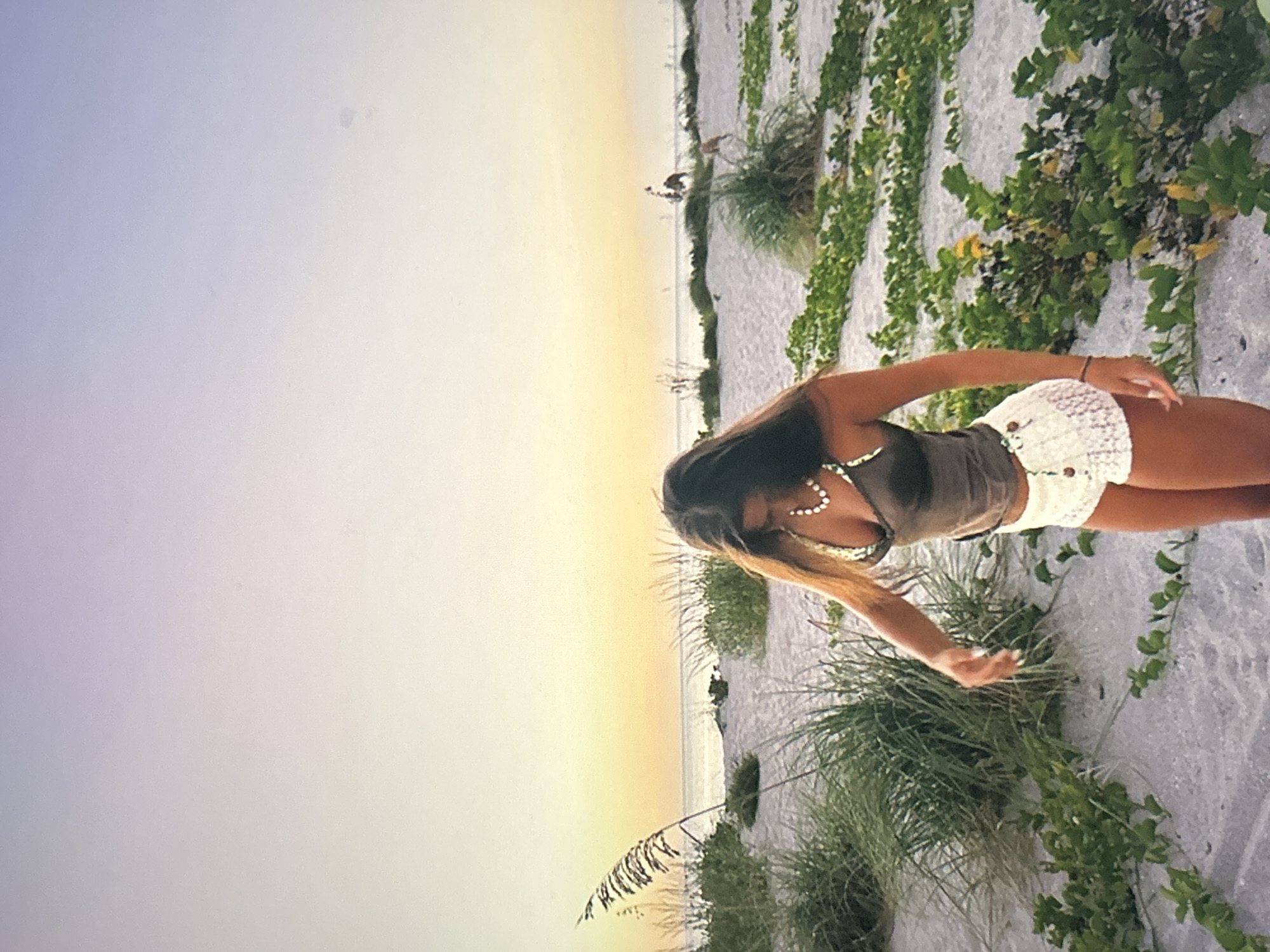 The image depicts a serene beach scene, likely during sunrise or sunset, with a person standing on the right side of the frame. The subject is a young woman with long, dark hair that has lighter highlights. She is dressed in a dark tank top and light-colored, possibly crocheted, shorts, complemented by a pearl necklace and earrings. Her body is gently bent forward, with her arms extended downwards, suggesting she is looking at or reaching for something on the sand.
The setting is a sandy beach with patches of green vegetation, including tall grasses resembling sea oats and sprawling, vine-like plants. In the distance, a calm body of water, likely the ocean or sea, meets the horizon. The sky exhibits a beautiful gradient, transitioning from soft, warm yellow and orange hues near the horizon to cooler, pale purple and blue tones overhead, indicative of dawn or dusk. Faint, dark figures, possibly birds or distant people, can be seen along the distant shoreline. The overall atmosphere of the image is peaceful and contemplative. No visible text is present in the media.