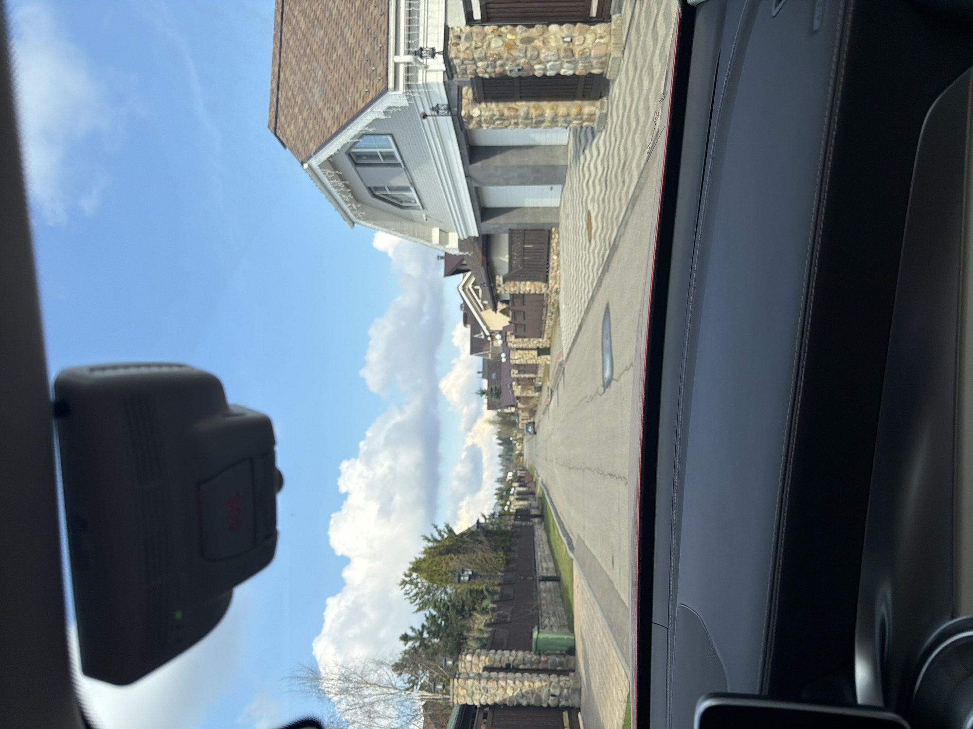 The image shows a residential street in Selyatino, Russia, viewed from inside a car. The street is lined with houses featuring stone facades and brown roofs. A sidewalk runs along the right side of the street. The sky is blue with white clouds. A car dashboard and a rearview mirror are visible in the foreground.