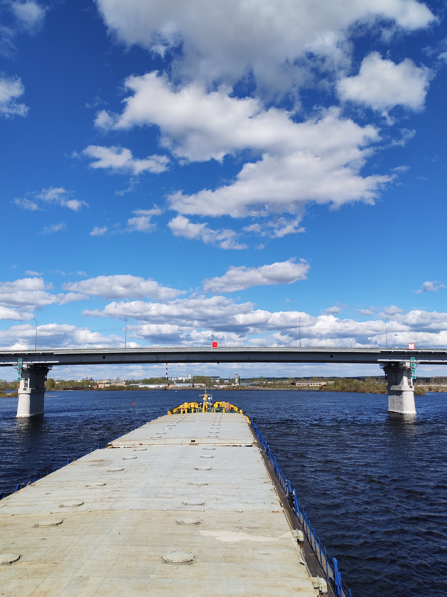 A full, eye-level shot shows the deck of a barge or pontoon, a body of water, a bridge, and the sky with scattered clouds on a bright, sunny day. The metal deck of the vessel is a uniform gray color, with a few rust spots. The deck contains numerous circular access ports. A blue rail runs along the edge of the barge. The dark blue water has some visible ripples. In the distance, a concrete bridge spans the water. The bridge pillars are a light gray color. A small patch of green trees lines the shore in the background. Beyond the trees, some low, indistinct buildings can be seen. The sky is a bright blue with scattered white, puffy clouds. The scene appears to be located in Sheksna, Russia, based on the general characteristics of the landscape.