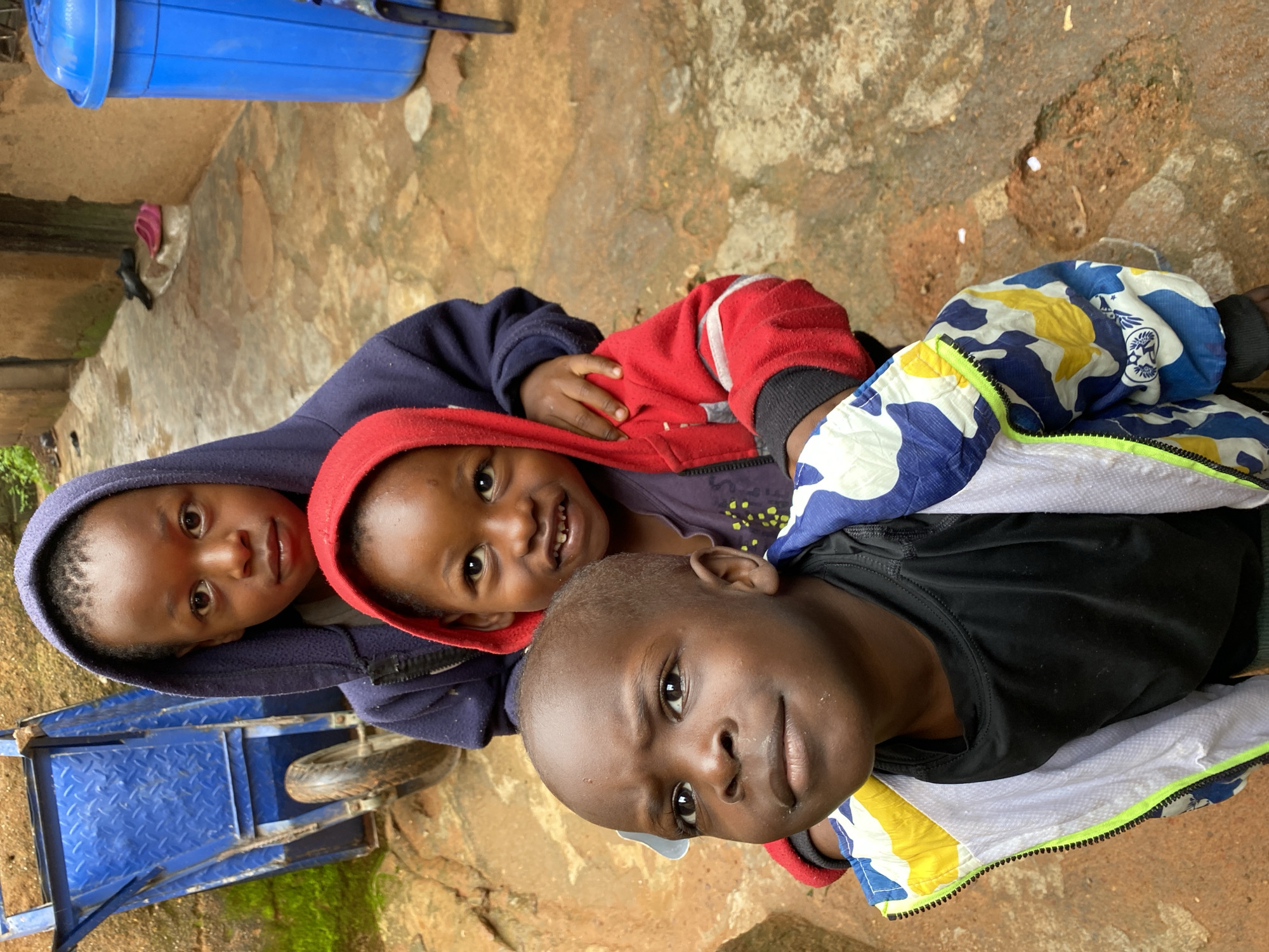 A close-up, eye-level, outdoor shot shows four young Black children standing in a line, their faces filling the frame. The children are wearing hooded jackets. The child in the front is wearing a black t-shirt and a multi-colored camouflage jacket with blue, yellow, and white patterns. The child behind them is wearing a red hooded jacket and a purple shirt. The third child, partially visible, is wearing a dark blue hooded jacket. The fourth child, the furthest back, is visible from the shoulders up, wearing a dark blue hooded jacket. The children are looking towards the camera. The background consists of a reddish-brown dirt pathway with scattered puddles, a tan-colored wall, and a bright blue trash bin on the right. A blue wheelbarrow is visible on the left side of the frame, next to the wall. The scene appears to be in a residential area within the city of Jos, Nigeria.