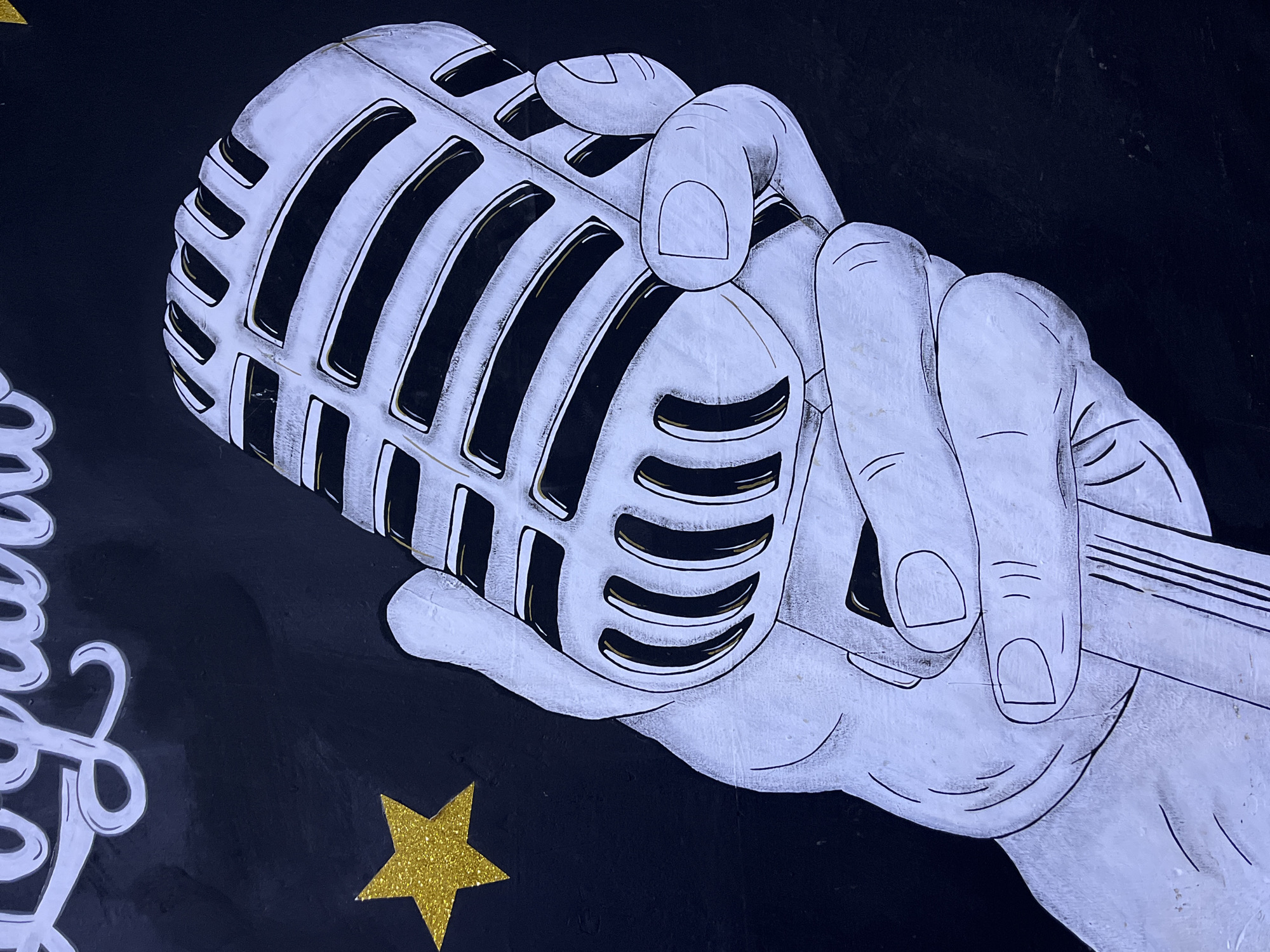 The image shows a mural on a black wall depicting a white gloved hand holding a vintage microphone. The microphone is silver with black grille openings and gold accents. A yellow glitter star is positioned to the left of the microphone. Faint white lettering, possibly part of a larger design, is visible at the top of the image. The mural appears to be located in Santa Ana, Venezuela.