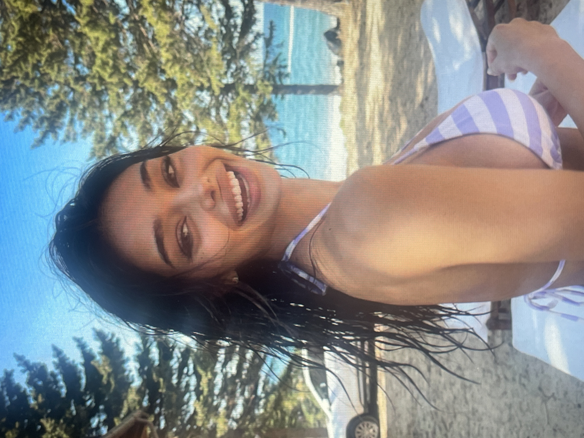 An image taken from a low angle shows a young woman with dark, wet hair, wearing a purple and white striped bikini, smiling at the camera. She is on a sandy beach, with trees and the ocean in the background. A white car is visible to the left. The scene appears to be a sunny day. The location in the city of Mantov, Czechia is not discernible from the image.