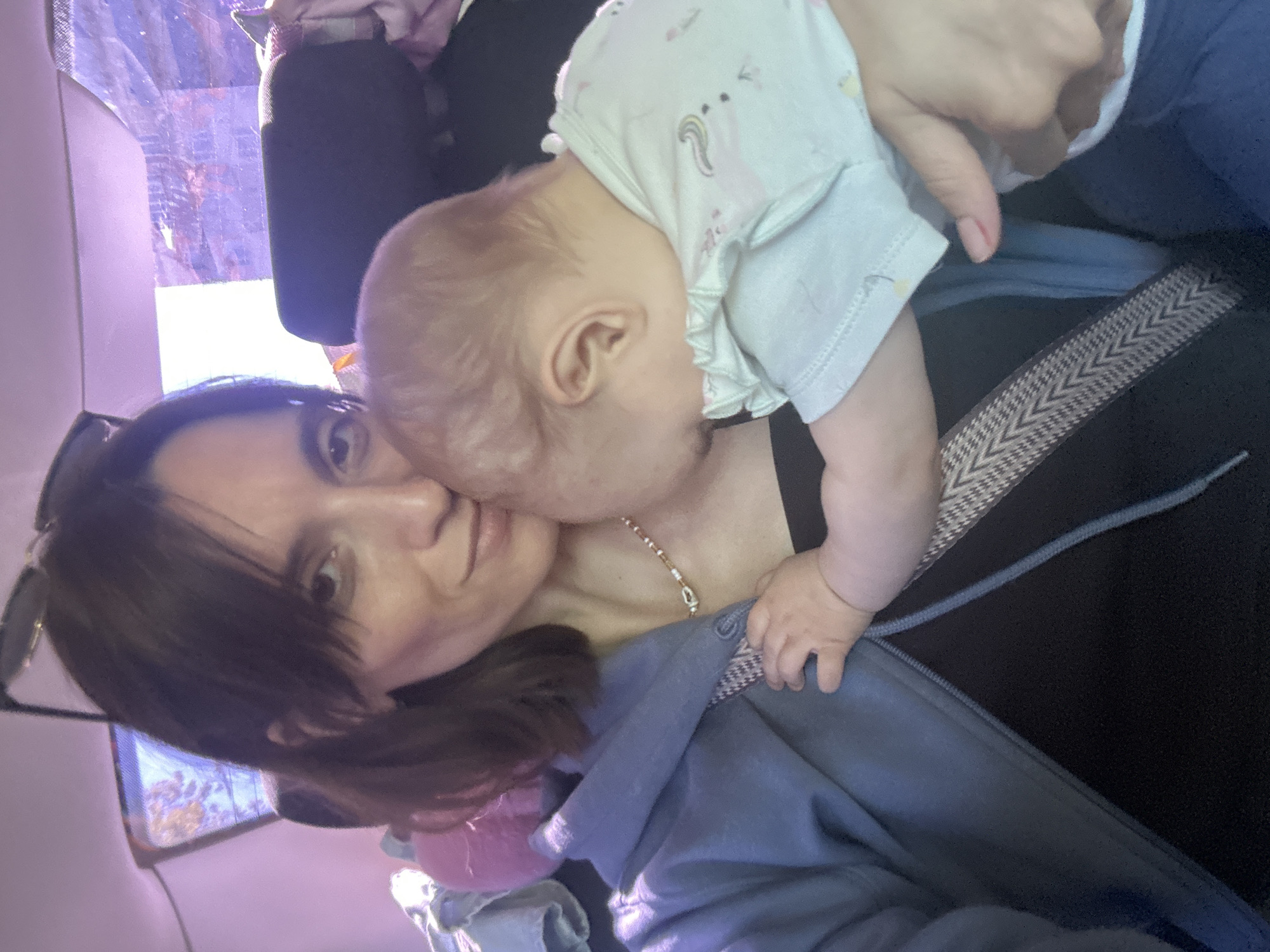 This image depicts a scene in the city of Moscow, Russia, featuring a female adult holding an infant within the interior of a vehicle. The adult female, positioned in what appears to be a rear passenger seat, has shoulder-length brown hair with bangs and wears sunglasses pushed onto her head. She is dressed in a light blue hooded sweatshirt over a black garment and a beaded necklace with a silver-toned clasp. Her right hand supports the infant while her left arm cradles the child. A patterned strap, likely a seatbelt or carrier strap with a black and white chevron design, crosses her body.
The infant, appearing to be under one year of age, has light, fine hair and is wearing a light-colored, short-sleeved top with a subtle unicorn and rainbow pattern and ruffled sleeves. The infant's face is turned towards and pressed against the adult's chest, obscuring facial features. One small hand rests on the adult's jacket.
The vehicle's interior shows a dark headrest behind the infant. Through the rear or side windows, bright daylight is visible, with a purple-tinted, somewhat obscured view of an outdoor environment, possibly urban structures, suggesting movement or a parked location during the day. A screen displaying an image is partially visible in the upper left background, and a pink, soft object rests behind the adult's left shoulder.