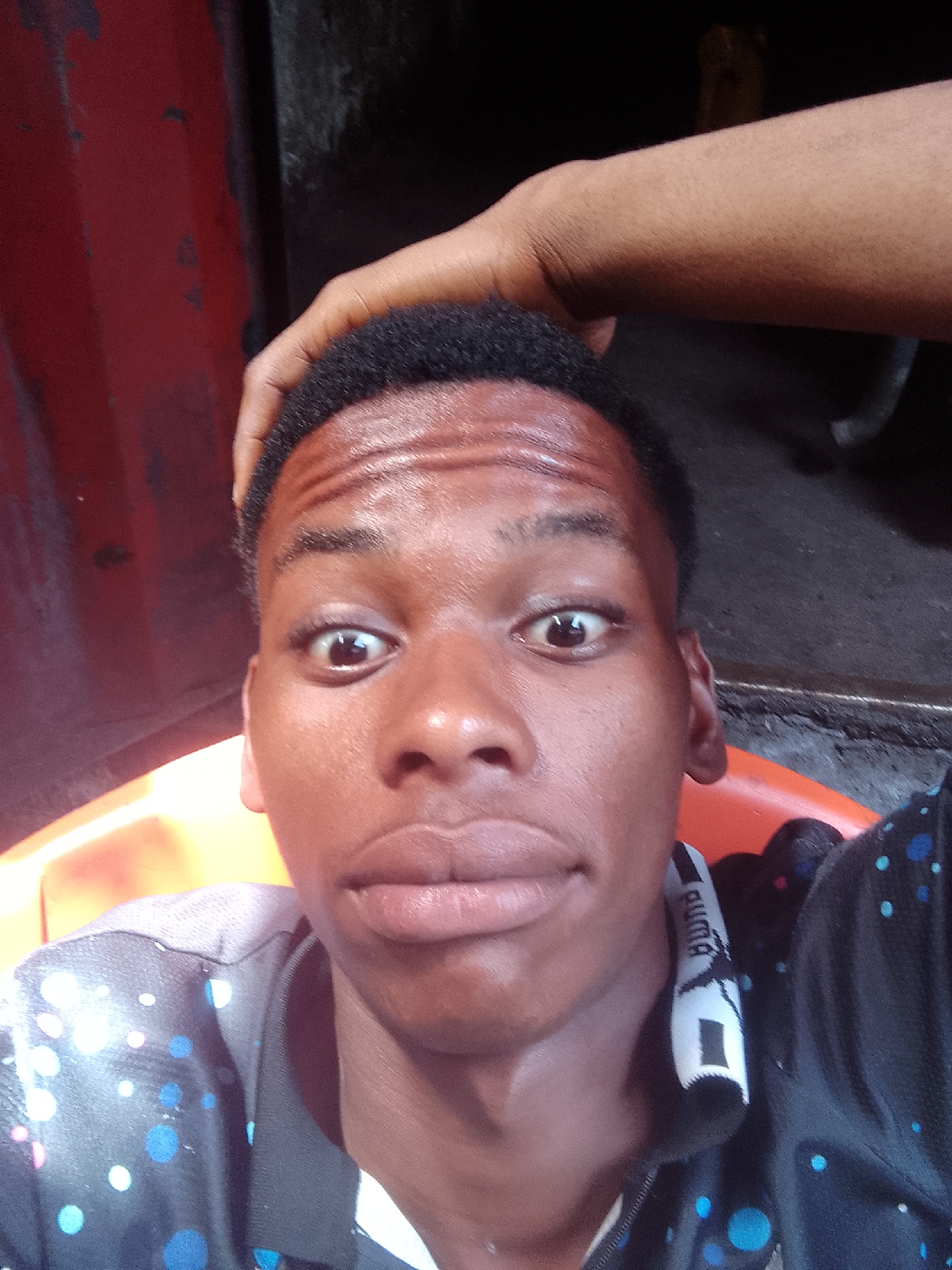 A selfie taken by a young Black man with dark hair and eyes, wearing a black and blue polka-dot jacket with a white Puma logo on the collar. His expression is wide-eyed with a slightly puckered mouth. A dark-skinned hand rests on his head. The background is blurred, but appears to be a vehicle or part of a building with red and grey hues. It is possible, based on the person's appearance and the environment, the photo was taken in the city of Onitsha, Nigeria.