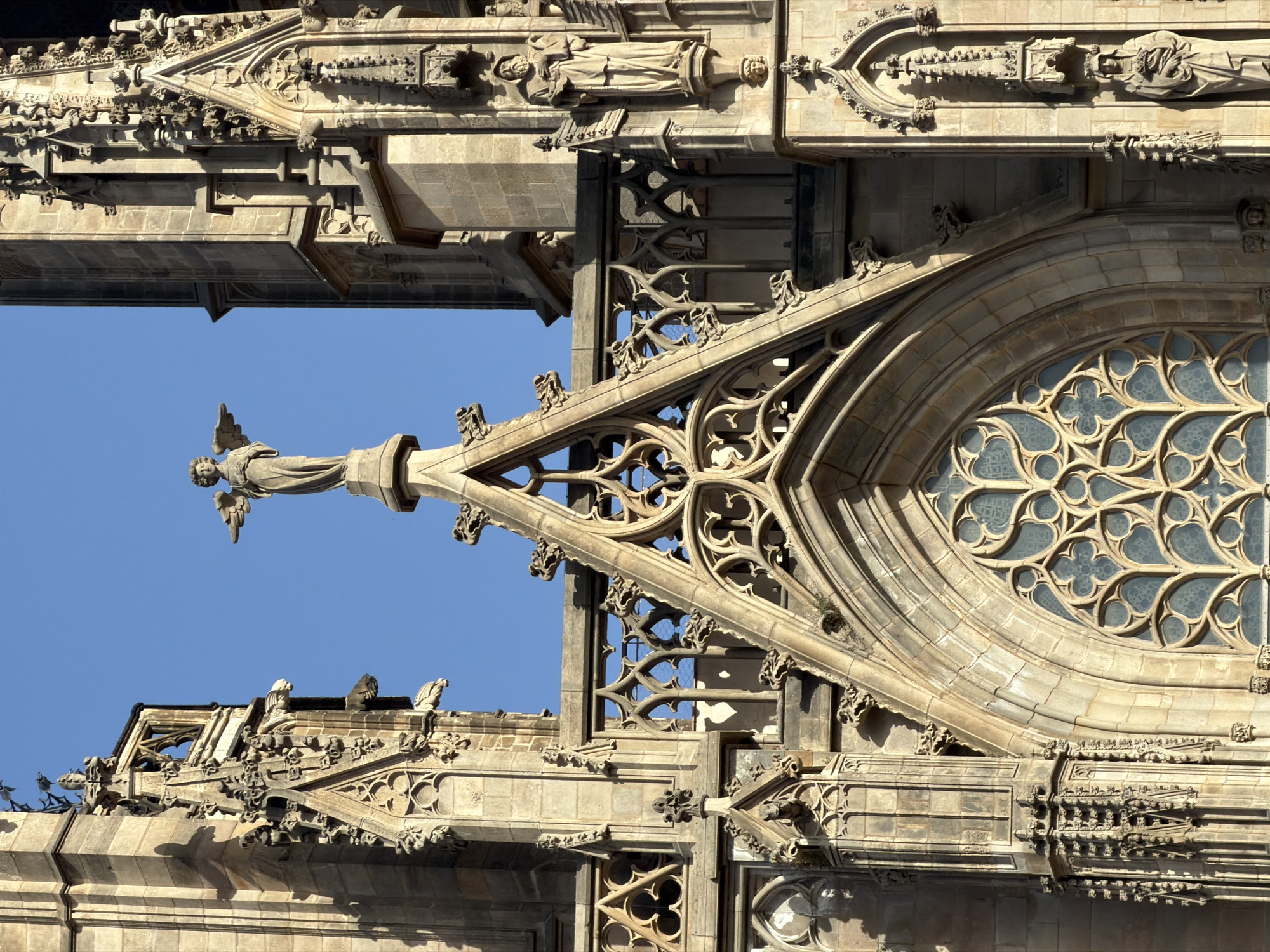 This is a close-up, low-angle, vertical shot of the exterior of the Barcelona Cathedral, located in the Gothic Quarter of Barcelona, Spain. The cathedral's facade, constructed from light-colored stone, features intricate Gothic architectural details. Prominently visible is an ornate, pointed archway framing a large rose window with a stylized, quatrefoil-like tracery. Above the archway, a statue of an angel with outstretched wings stands on a pedestal. The surrounding stonework is adorned with gargoyles, decorative carvings, and niche statues, including one of a robed male figure holding an object. The sky above is a clear, bright blue.