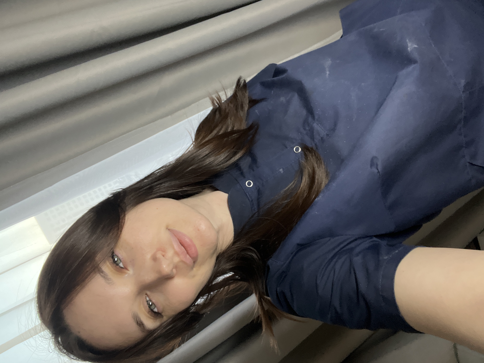 A self-portrait captures a young woman from the chest up, positioned diagonally within the frame. She has long, wavy dark brown hair cascading around her, and striking light blue or grey eyes. Her gaze is directed calmly towards the camera, and her expression is serene with a slight, natural smile on her lips.
She is dressed in a dark navy blue, long-sleeved top, possibly a shirt or light jacket, which features silver snap buttons visible on the placket near her right shoulder. The fabric of her garment shows some light-colored marks or lint.
The setting appears to be indoors in Tyumen, Russia, likely a room with a window. In the upper left and behind her head, light-colored, possibly grey, pleated curtains or blinds are visible, suggesting natural light from a window illuminates her face. A white ledge or frame is discernible below the curtains. The background behind her body is darker and less defined, contributing to the indoor ambiance. The time of day appears to be during daylight hours. No text is visible in the image.