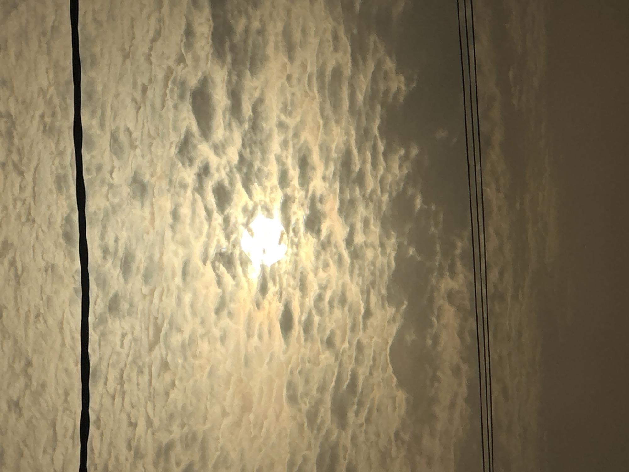 The image depicts a sky with a dense layer of altocumulus clouds, partially obscuring the sun, creating a diffused light effect. Electrical wires are visible in the left and right side of the frame. The low angle perspective suggests the photo was taken at ground level. The light indicates late afternoon or early morning. There is no evidence to support it being taken in the city of Bukuru, Nigeria.