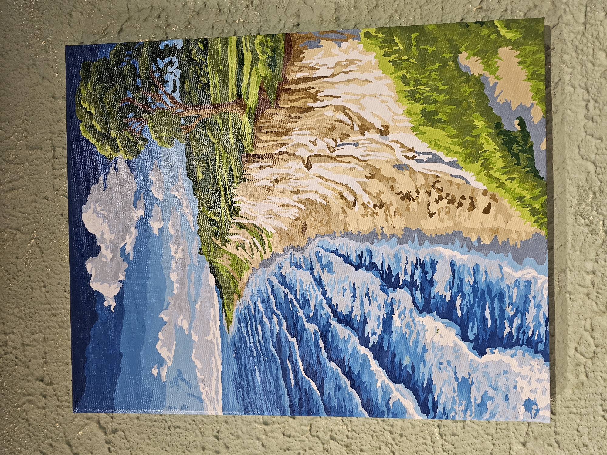 A painting depicts a coastal cliff landscape. The scene features a body of water with waves on the left, a sandy beach, and a steep, light-colored cliff face. Above the cliff, there is green grassy terrain with a large tree on the right side of the canvas. The sky above is blue with white clouds. The painting is framed and displayed on a textured, greenish-gray wall.