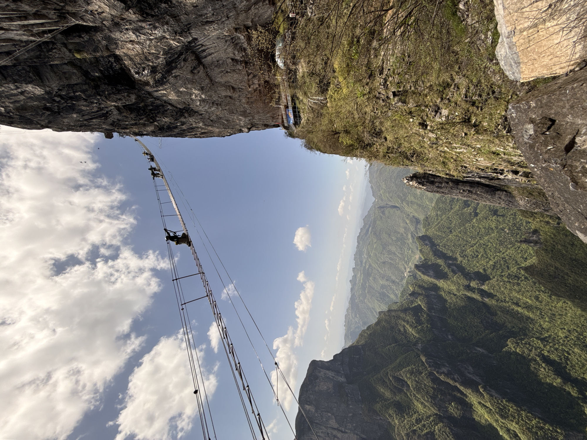 The image shows a section of the Via Ferrata in the city of Yongding District, China. The scene depicts a challenging climbing route constructed on a steep rock face. The route consists of a series of metal rungs and cables anchored into the rock. Several climbers wearing safety harnesses and helmets are ascending the Via Ferrata. They are clipped onto the cables, which serve as a safety measure in case of a fall. The environment surrounding the Via Ferrata features a rugged landscape with lush green vegetation covering the mountains. The sky is clear with scattered clouds.