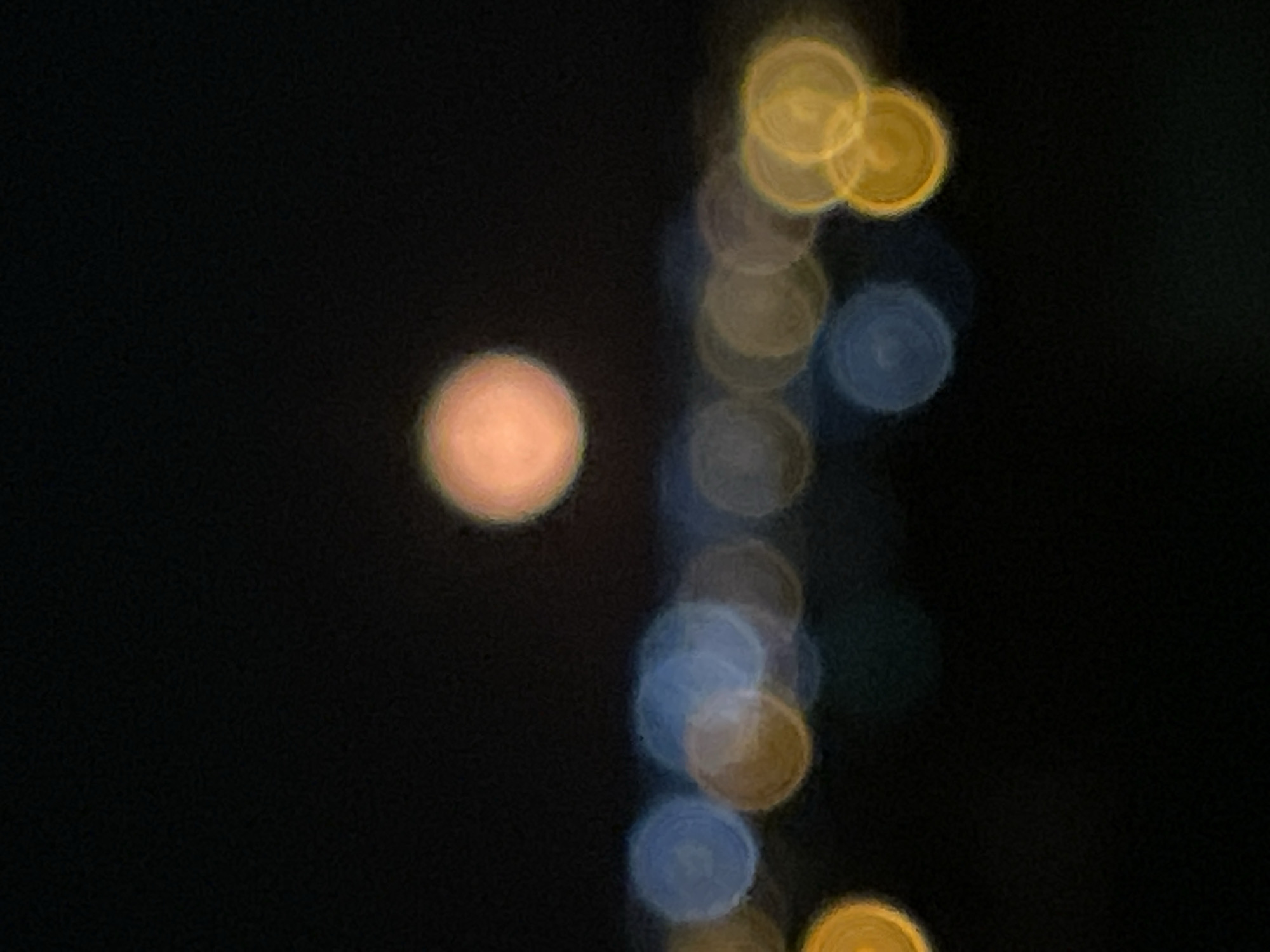 The image is a blurry, out-of-focus photograph taken at night. On the left side of the image, a single, large, soft orange-colored circular shape is visible. To the right, a vertical line of smaller, overlapping blurry circles extends from the top to the bottom of the frame. These circles alternate in color, appearing yellow, blue, and grey. The background is entirely dark, obscuring any further details about the environment. There is no discernible information in the image to definitively locate it in Rojales, Spain or to identify any people, subjects, objects, actions, or interactions.