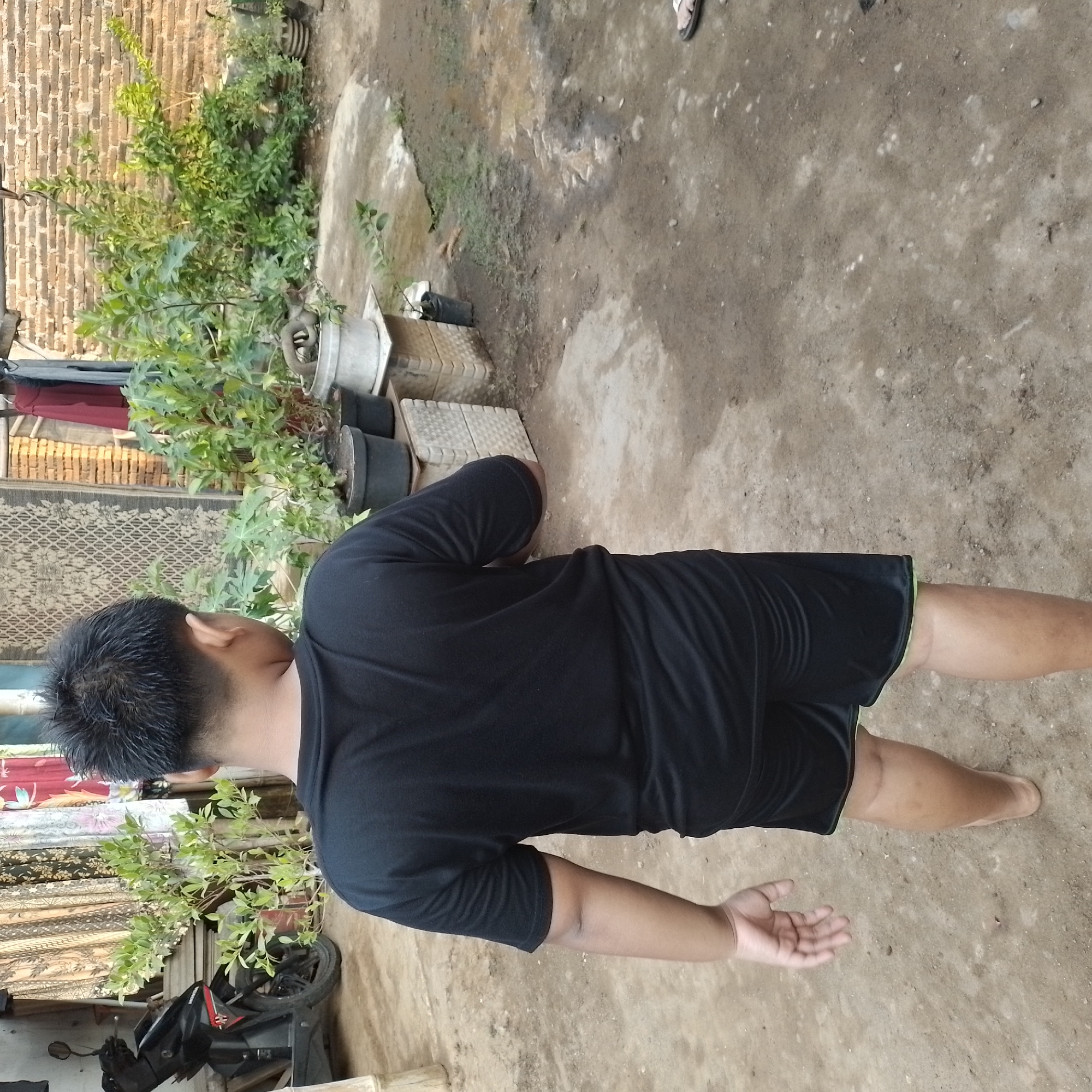 The image shows a young boy from behind, wearing a black t-shirt and black shorts, standing in an outdoor area. He is barefoot. To his left, a black motorcycle is parked. Behind him, there are several potted plants and some hanging fabrics. A brick wall is visible in the background, suggesting an urban or semi-urban setting. The ground is dirt and gravel. The scene is likely a residential yard or alleyway. Based on the architectural style and vegetation, it is plausible this scene is located in Jatisobo, Indonesia.