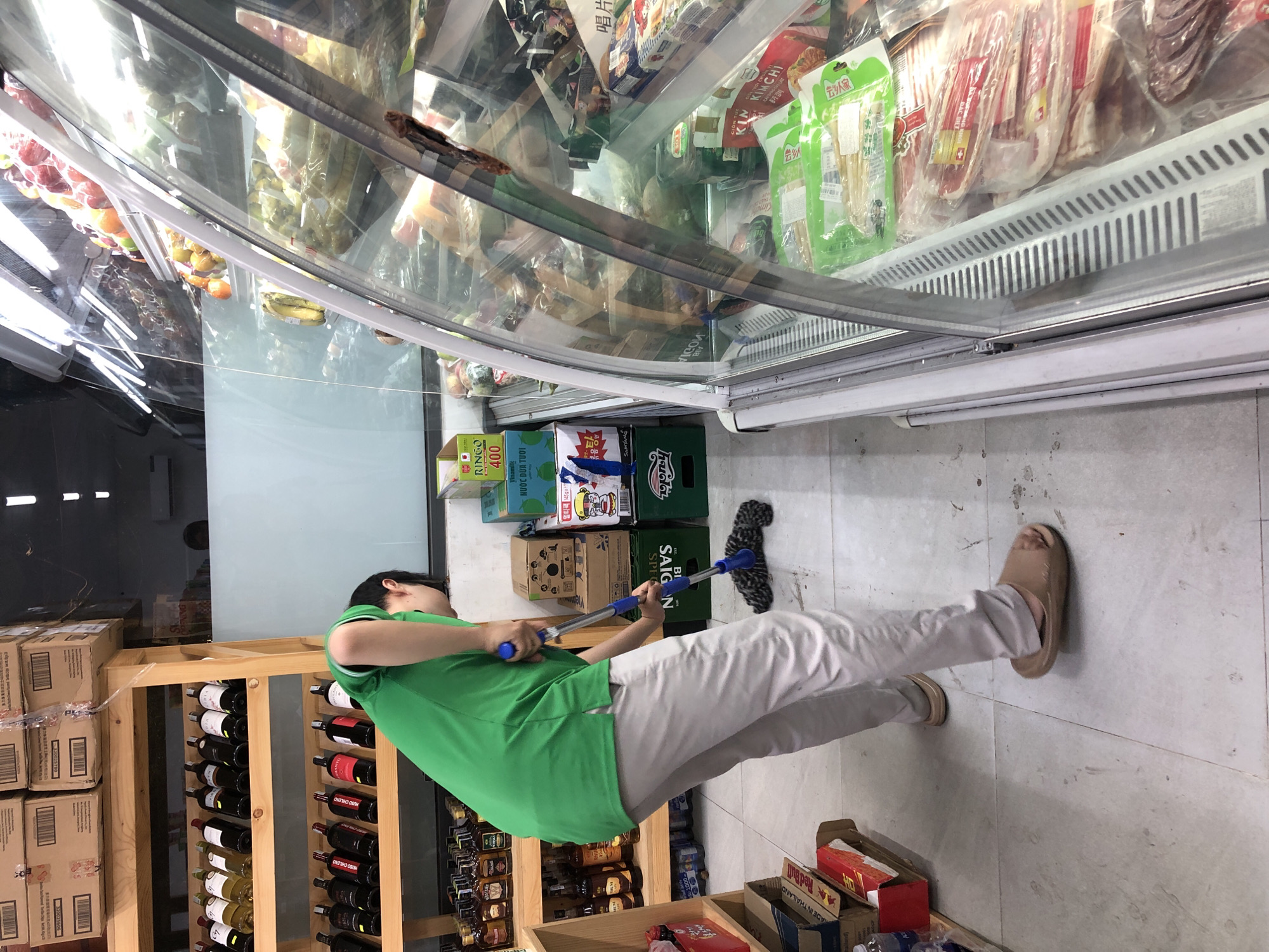 A person in a green t-shirt and light-colored pants is cleaning the floor with a mop in what appears to be a convenience store or a small market. The store is filled with shelves of products, including bottled beverages, boxes, and items in a refrigerated display case. The refrigerated case contains various food items, possibly meats and produce, visible through its glass doors. Next to the person, there are shelves stocked with wine bottles. In the background, more shelves are filled with boxed goods and drinks. The floor is tiled and looks somewhat dusty, indicating the need for cleaning. The lighting suggests it could be daytime, as there are bright lights from the store's ceiling and the refrigerated display. Visible text includes "Red Bull," "SAIGON," "Vinamilk," "NƯỚC DỪA TƯƠI," "KIN KIMCHI," and various Korean product labels. The overall scene depicts a routine cleaning task in a retail environment.