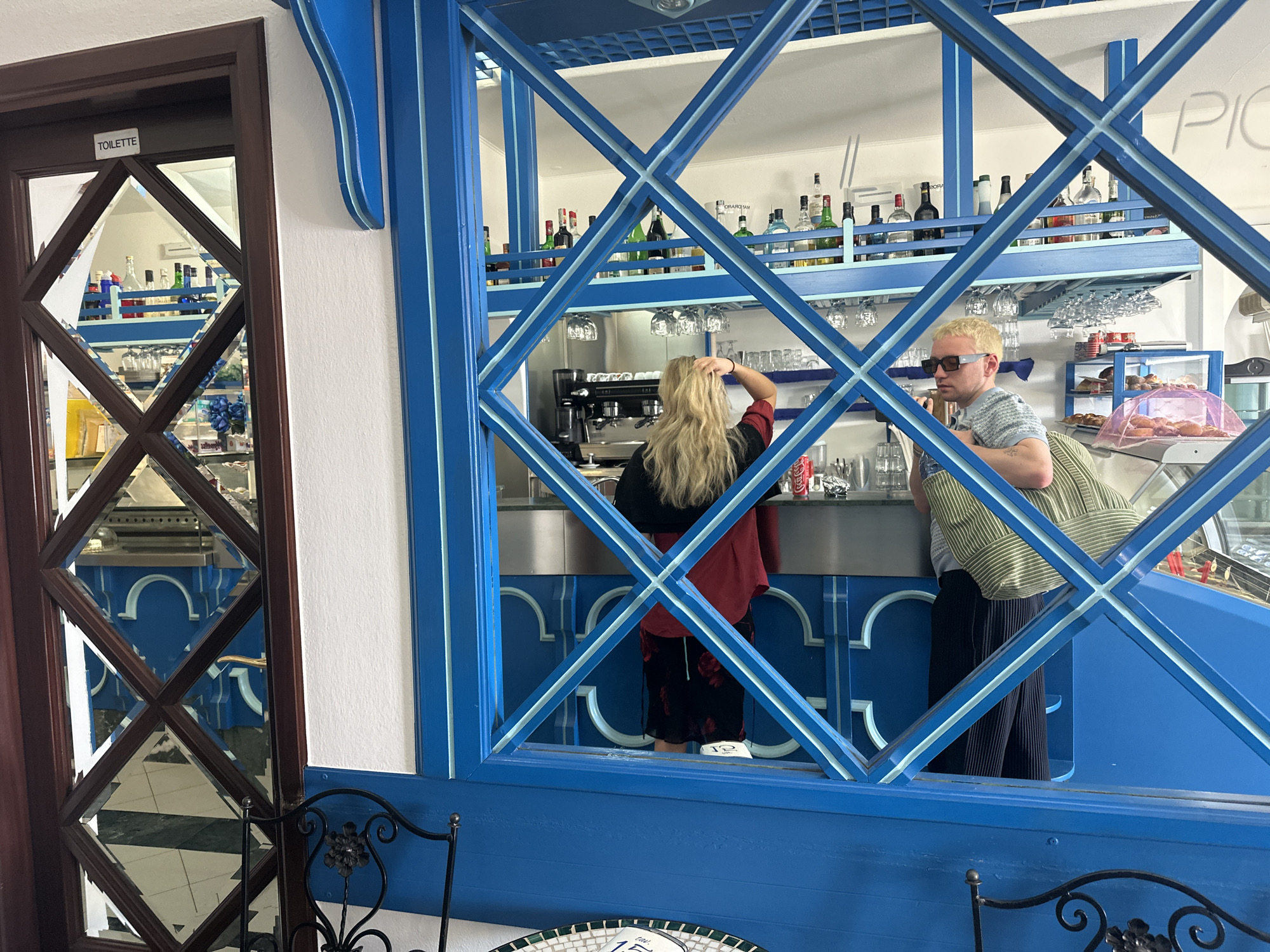 The image presents an indoor scene within a cafe or bar, viewed through a decorative blue wooden lattice screen with lighter blue stripe accents, which acts as a prominent foreground element.
Two individuals are visible behind the lattice, standing at a service counter. On the left, a woman with long, light blonde hair is seen from behind, wearing a black top with a red or maroon layer. Her right hand is raised, touching her head. To her right stands a man with short, bleached blonde hair, wearing sunglasses, a striped t-shirt, and dark pleated trousers. He carries a striped shoulder bag. Both appear to be patrons or staff interacting at the counter.
The bar area features shelves lined with various liquor bottles and an espresso machine. Wine glasses hang upside down from racks below the shelves. To the right of the man, a glass display case holds an assortment of pastries or baked goods. The counter below is adorned with blue panels featuring decorative arch cutouts. The overall decor uses a distinct blue and white color palette, suggesting a Mediterranean influence.
On the far left, a dark brown door with diamond-shaped mirrored panels is visible. A white sign reading "TOILETTE" is affixed to the top of this door. The mirrored panels reflect parts of the interior, including a black and white checkered floor. Partially visible on the white wall above the bar are light-colored letters that seem to spell "PIC" or "PICC", possibly part of the establishment's name. The bright lighting suggests it is daytime. The setting, with its blue and white theme and context of Lipari, Italy, evokes a coastal European ambiance.