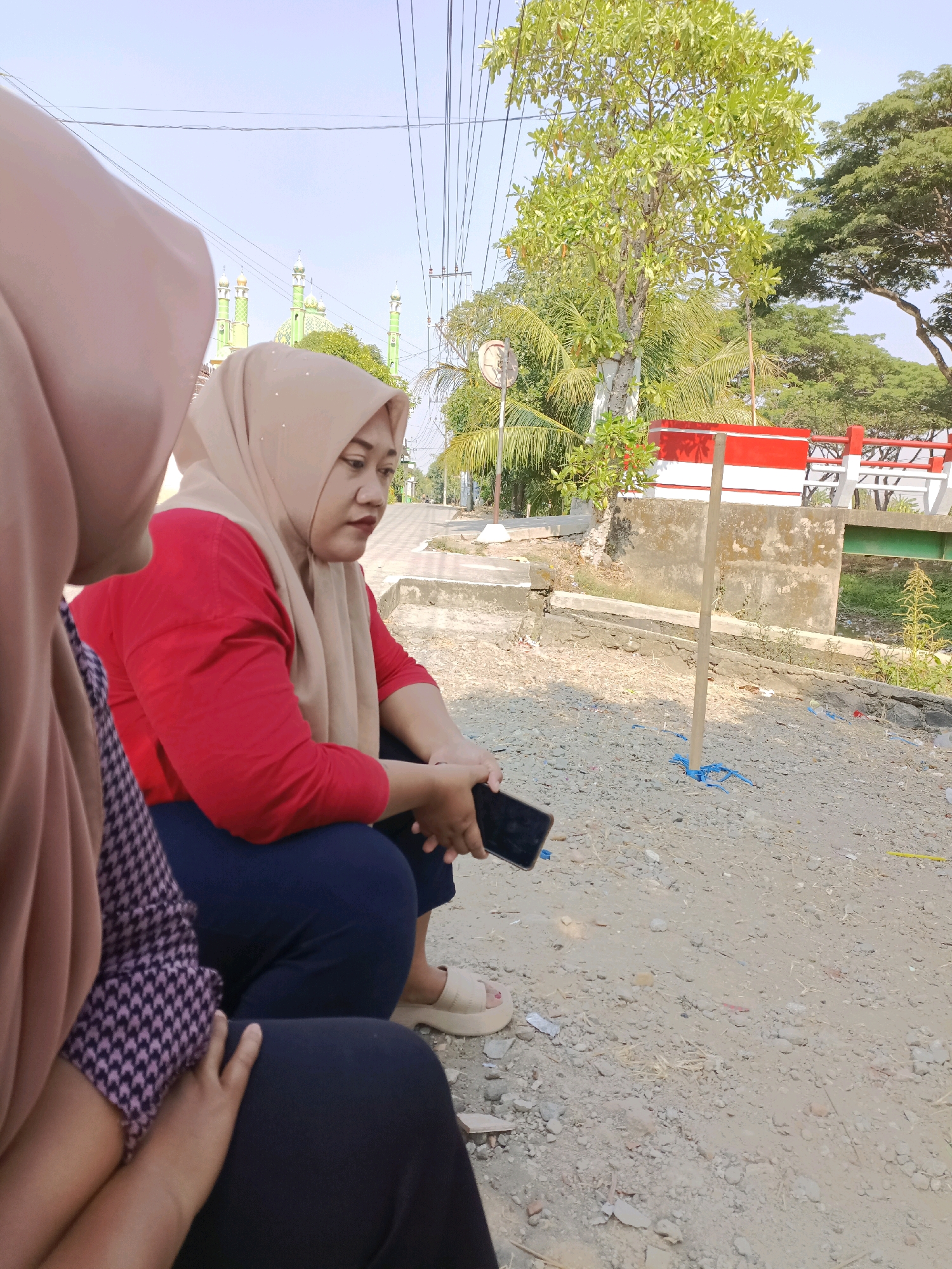 Two women are sitting on the ground in what appears to be an outdoor public space, possibly a park or roadside area, in Cangkring Rembang, Indonesia. The woman in the foreground on the right wears a light brown hijab and a red long-sleeved shirt with dark blue pants. She is looking down and to her right, with her hands clasped in her lap. She is holding a black smartphone in her right hand. The woman on the left, partially visible, wears a pink hijab and a patterned long-sleeved garment. Her left hand rests on the thigh of the woman on the right.
The environment is characterized by a dirt and gravel ground. In the background, a road with a patterned pavement is visible, leading towards a distant green mosque with multiple domes and minarets. Trees with green foliage are scattered throughout the scene, and power lines are strung across the sky. A red and white striped barrier or wall is also present on the right side of the image. The overall lighting suggests it is daytime, with clear sky.