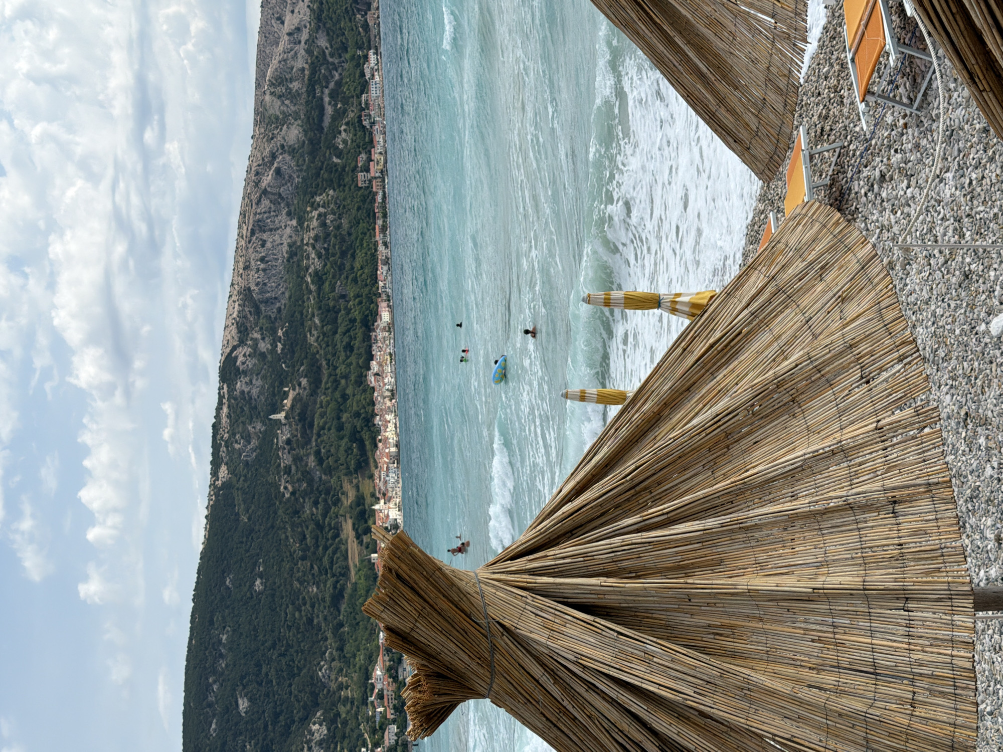 This is a medium shot of a beach scene in Općina Baška, Croatia, on a cloudy day. In the foreground, two thatched umbrellas partially obscure a pebble beach with orange lounge chairs. The umbrellas are made of dried reeds tied together with string. The beach transitions into a turquoise sea with moderate waves. Several people are visible in the water; one person is on an inflatable ring, while others appear to be swimming or floating. In the background, a town with white and red-roofed buildings lines the coast, backed by a densely forested, rocky hillside. A small white tower is visible on the hillside. The sky is filled with scattered white clouds.