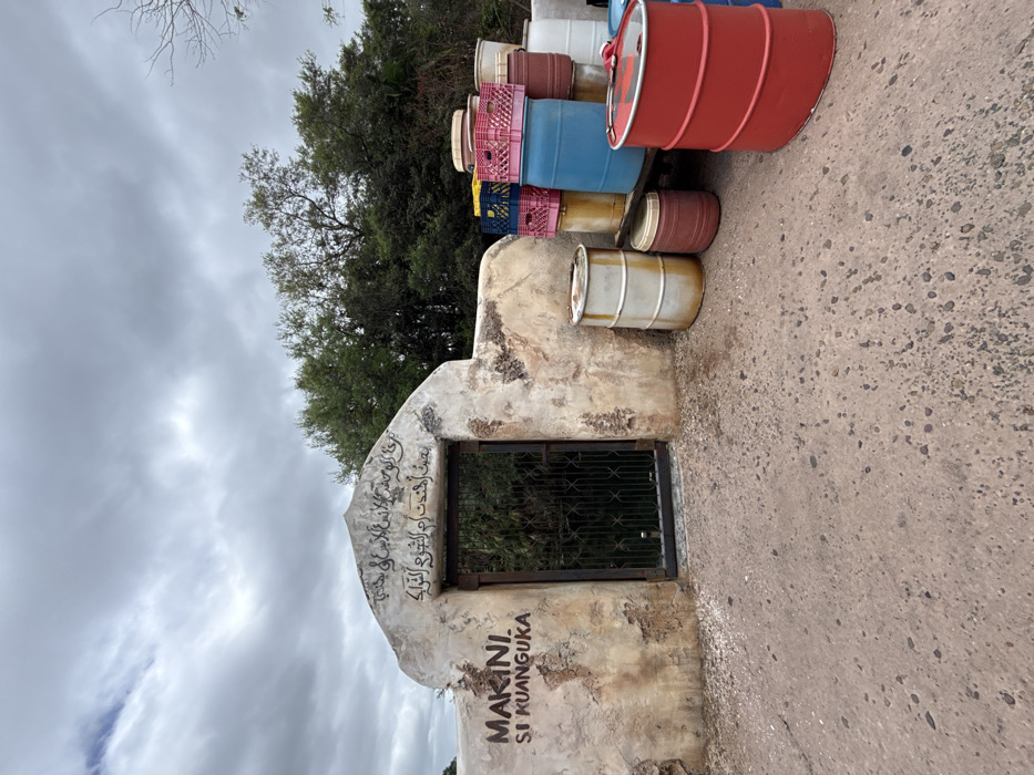 The scene depicts an outdoor area in Bay Lake, United States, featuring a weathered stone archway with an iron gate and various storage containers. The archway, light in color with visible erosion and dark markings, contains a closed, dark metal gate with vertical bars. Above the gate, black Arabic script is painted, and to the left, the words "MAKINI. SI KUANGUKA" are painted in black. To the right of the archway, an assortment of plastic and metal barrels, along with plastic crates, are stacked. Visible barrels include red, blue, white, and yellow plastic types, alongside at least one white metal barrel with rust, and a prominent red metal barrel in the foreground. Plastic crates, primarily pink, blue, and yellow, are stacked on top of some barrels. The ground is a rough, light brown or reddish-brown paved surface with embedded small stones. In the background, dense green trees and foliage are visible under an overcast, gray sky. No individuals are present.